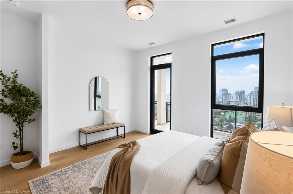 Staged bedroom with a white bed, tan pillows, and city views through tall black-framed windows.