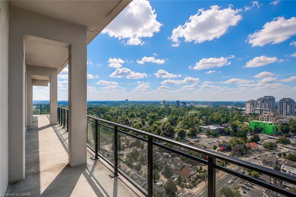 Condo balcony overlooking a leafy neighborhood and distant buildings, with glass railings and concrete columns.