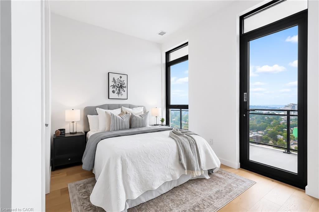 Staged high-rise bedroom with a large bed and tall windows overlooking trees and nearby buildings.