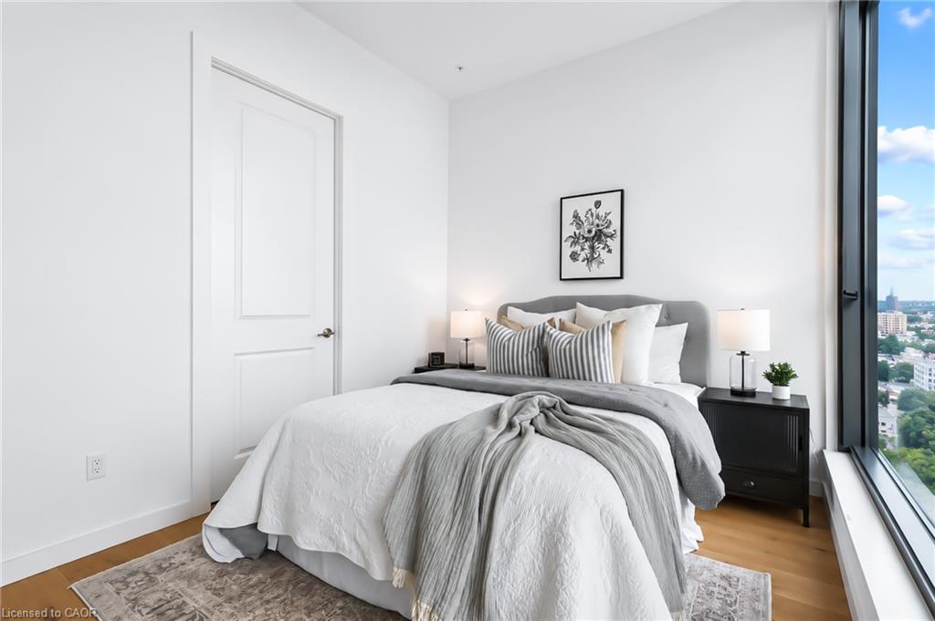 Staged condo bedroom with a white bed, gray headboard, and floor-to-ceiling window overlooking the city.