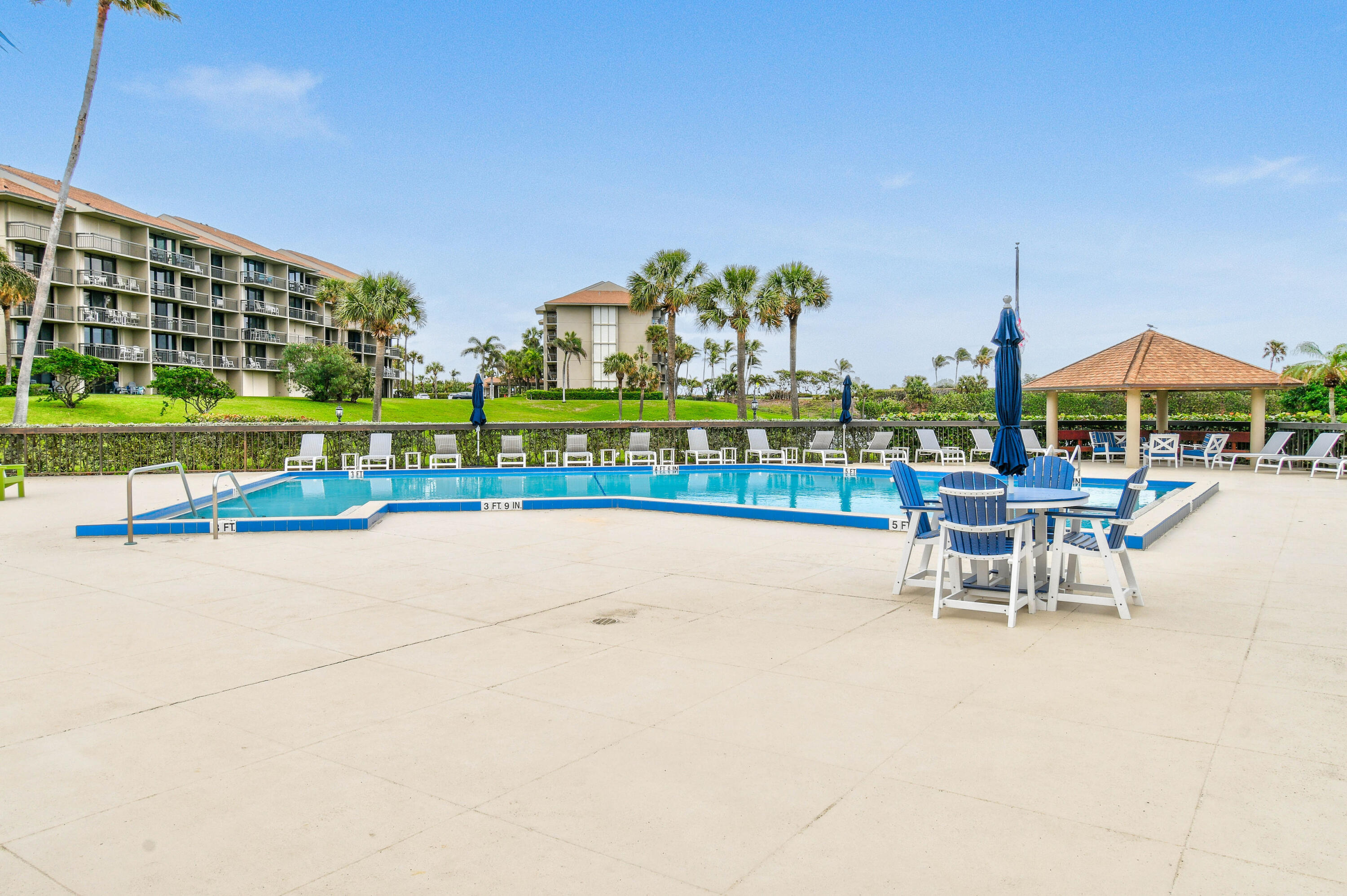 Outdoor condominium pool with palm trees, rows of lounge chairs, and a covered gazebo.