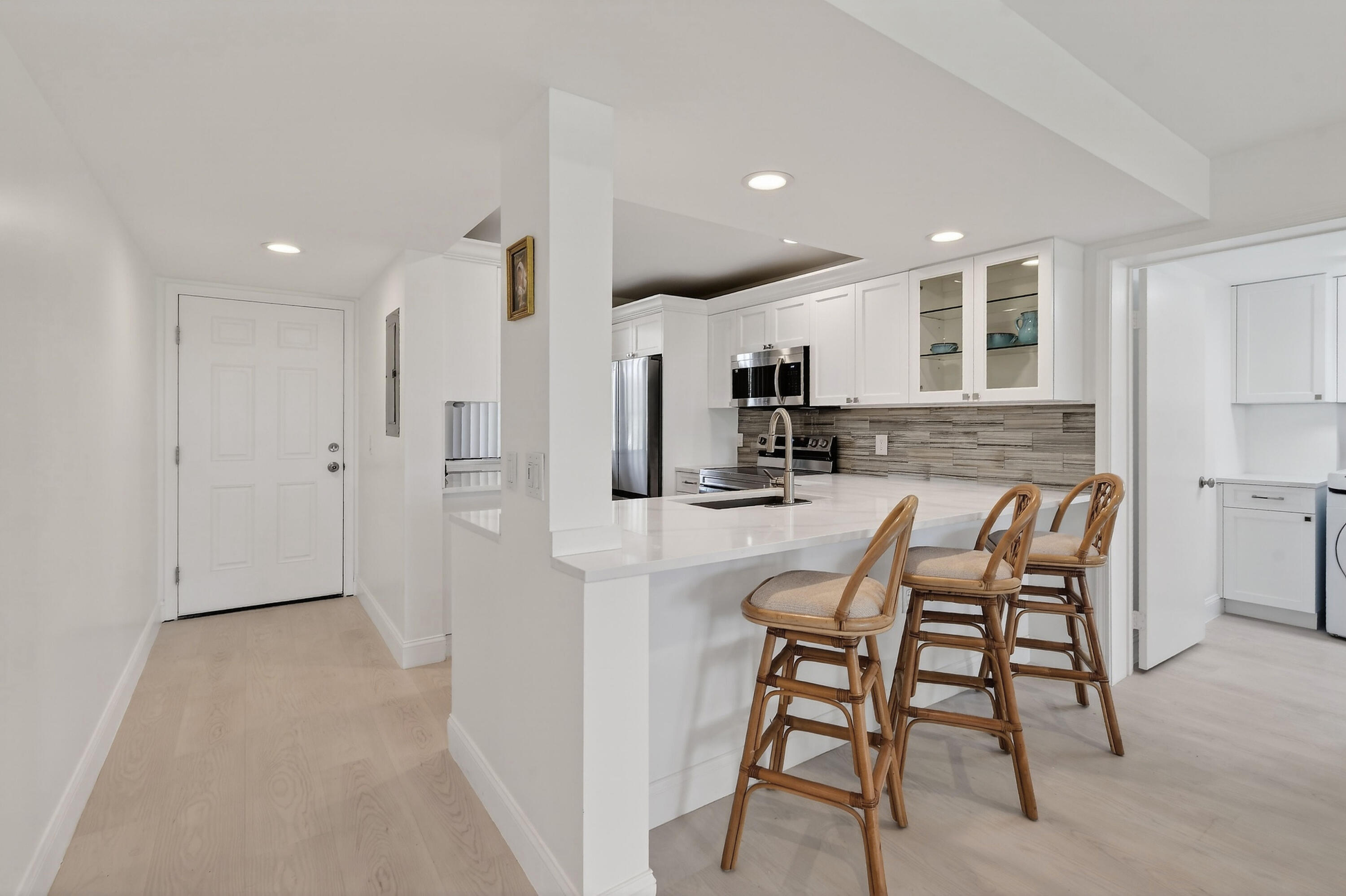 Bright white kitchen with stainless appliances, gray tile backsplash, and a peninsula with three wooden stools.