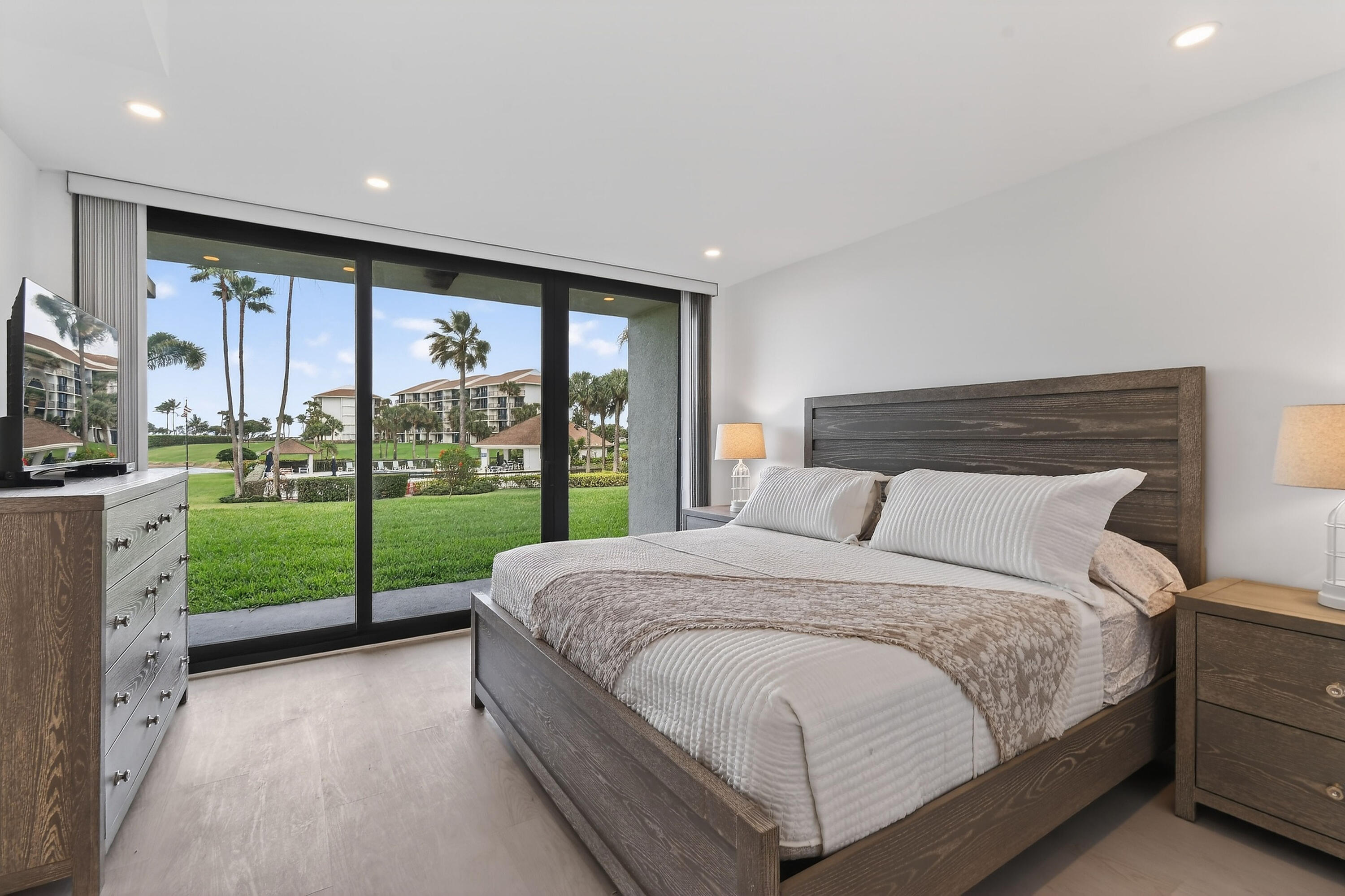 Furnished bedroom with sliding glass doors overlooking a palm-lined lawn and neighboring buildings.