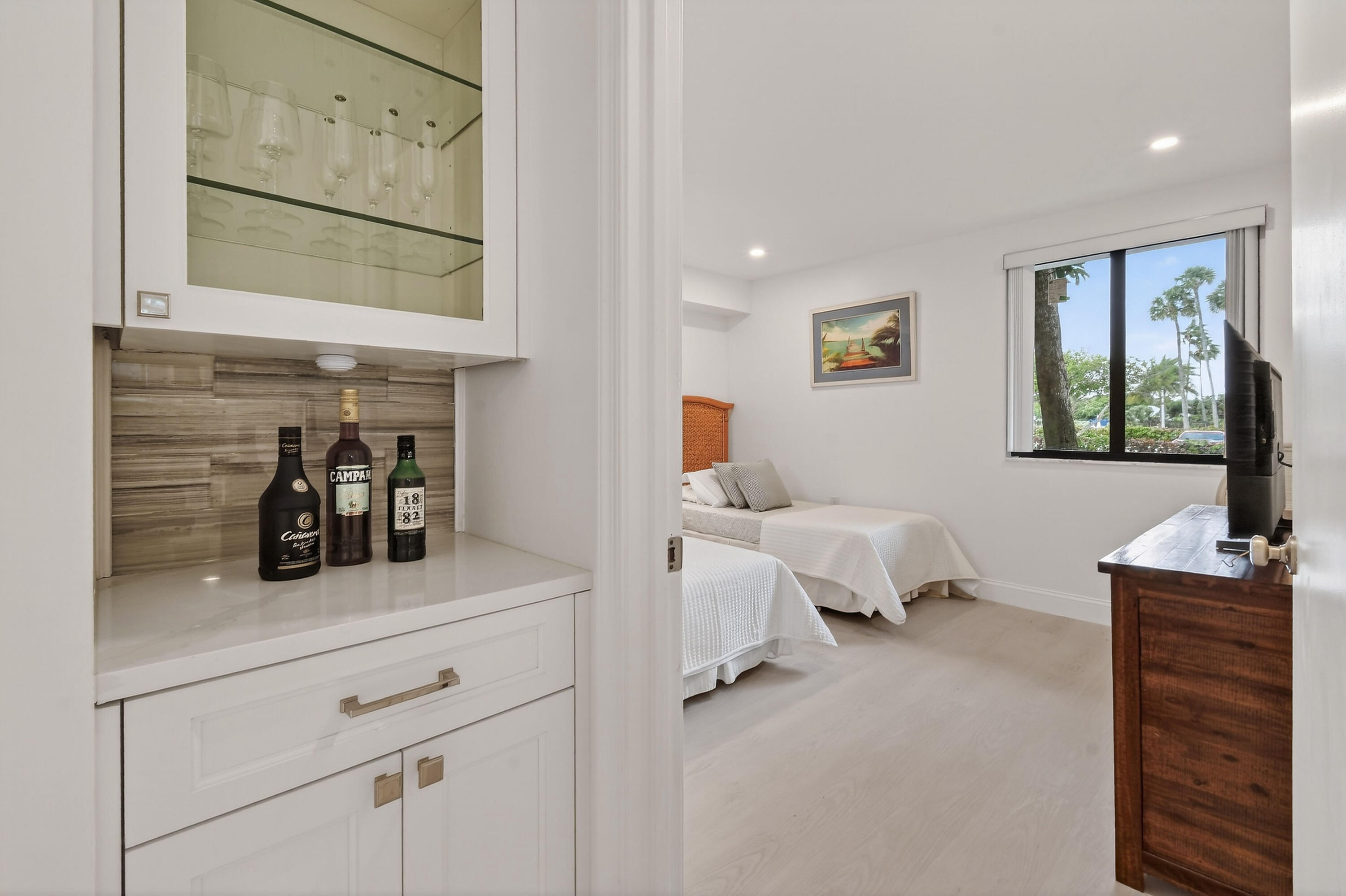 Bedroom with two twin beds and a built-in beverage cabinet beside a window overlooking palm trees.