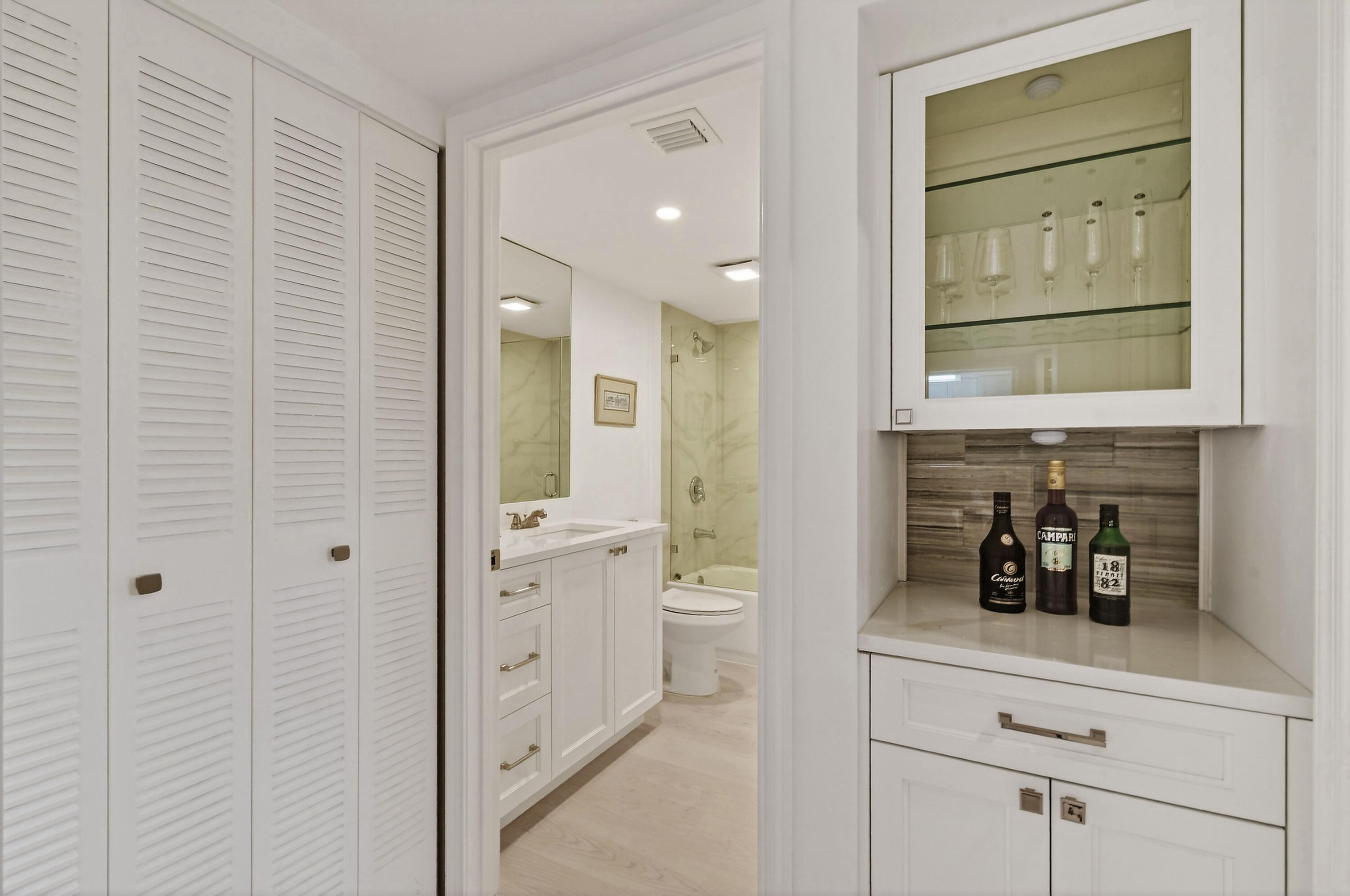 Bathroom with white vanity and glass shower beside a built-in cabinet with glassware and bottles.