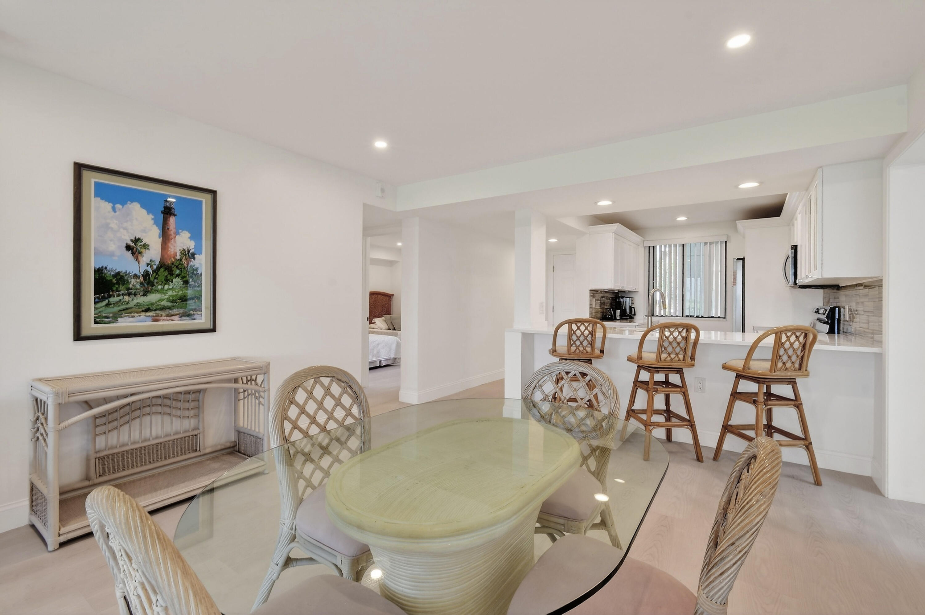 Dining area with a glass-topped wicker table opening onto a white kitchen and breakfast bar.
