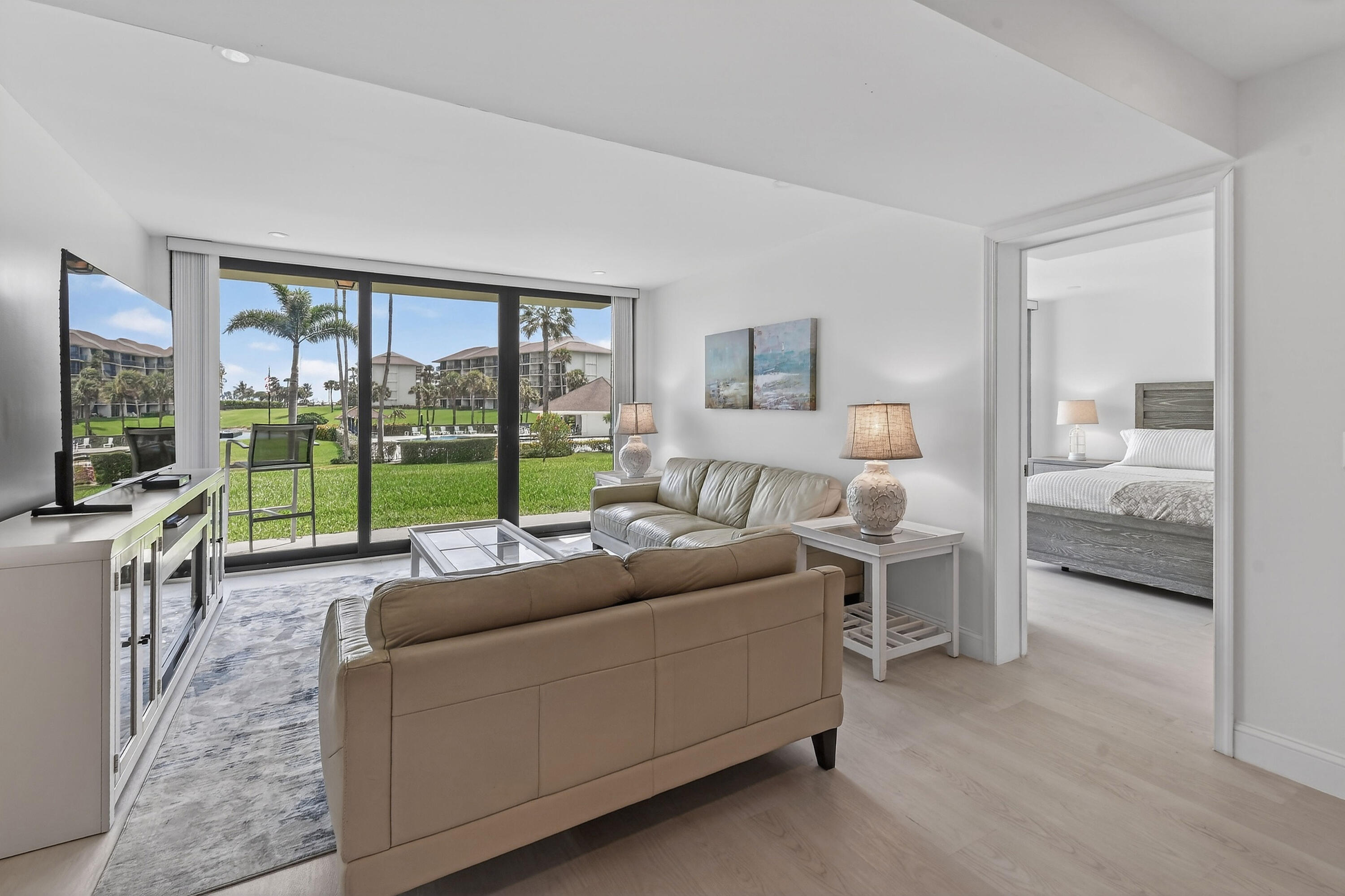 Furnished condo living room with sliding glass doors overlooking a palm-lined lawn; bedroom visible through doorway.