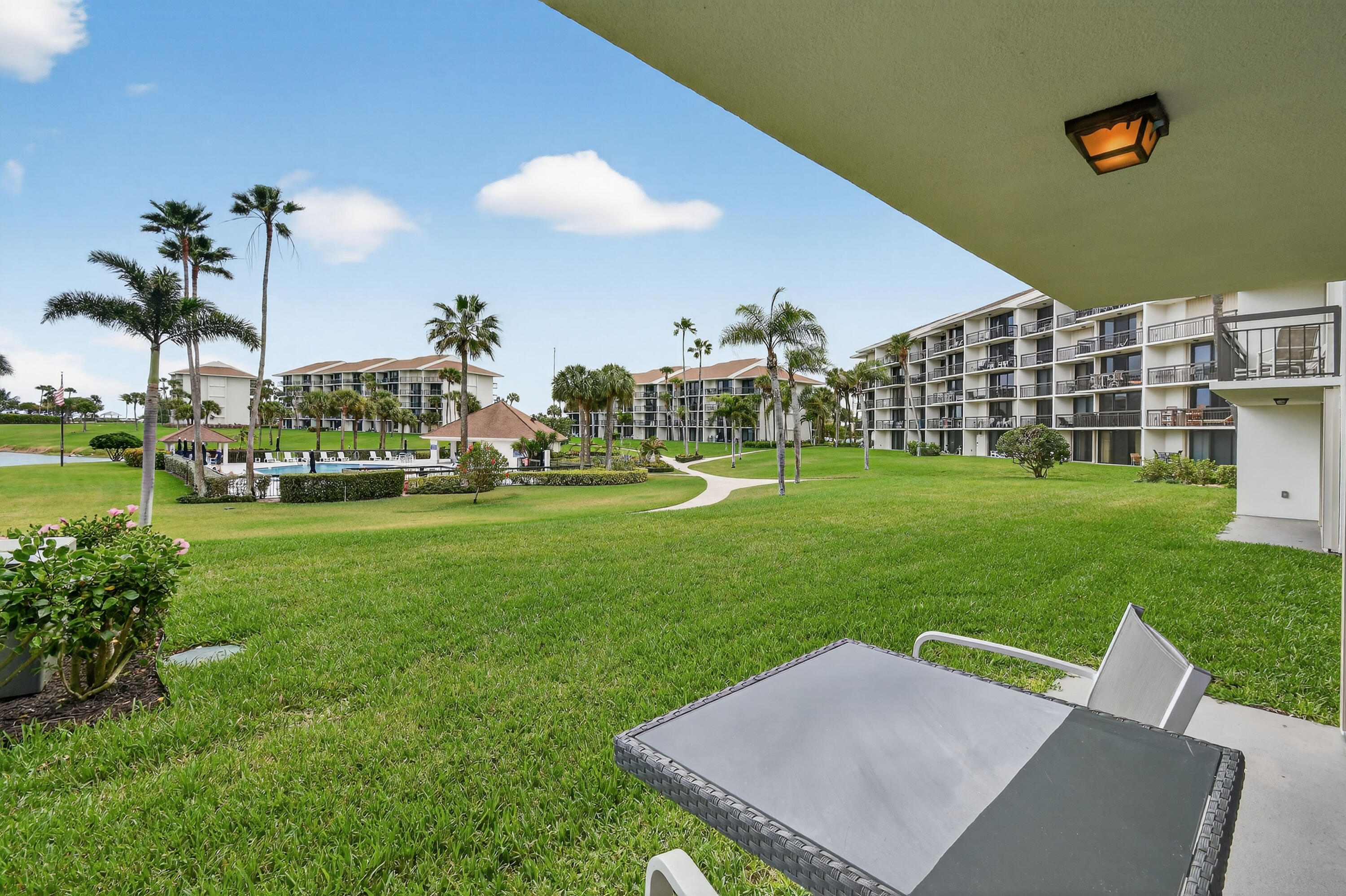 Covered condo patio overlooking palm-lined grounds, a curving walkway, and a fenced pool.