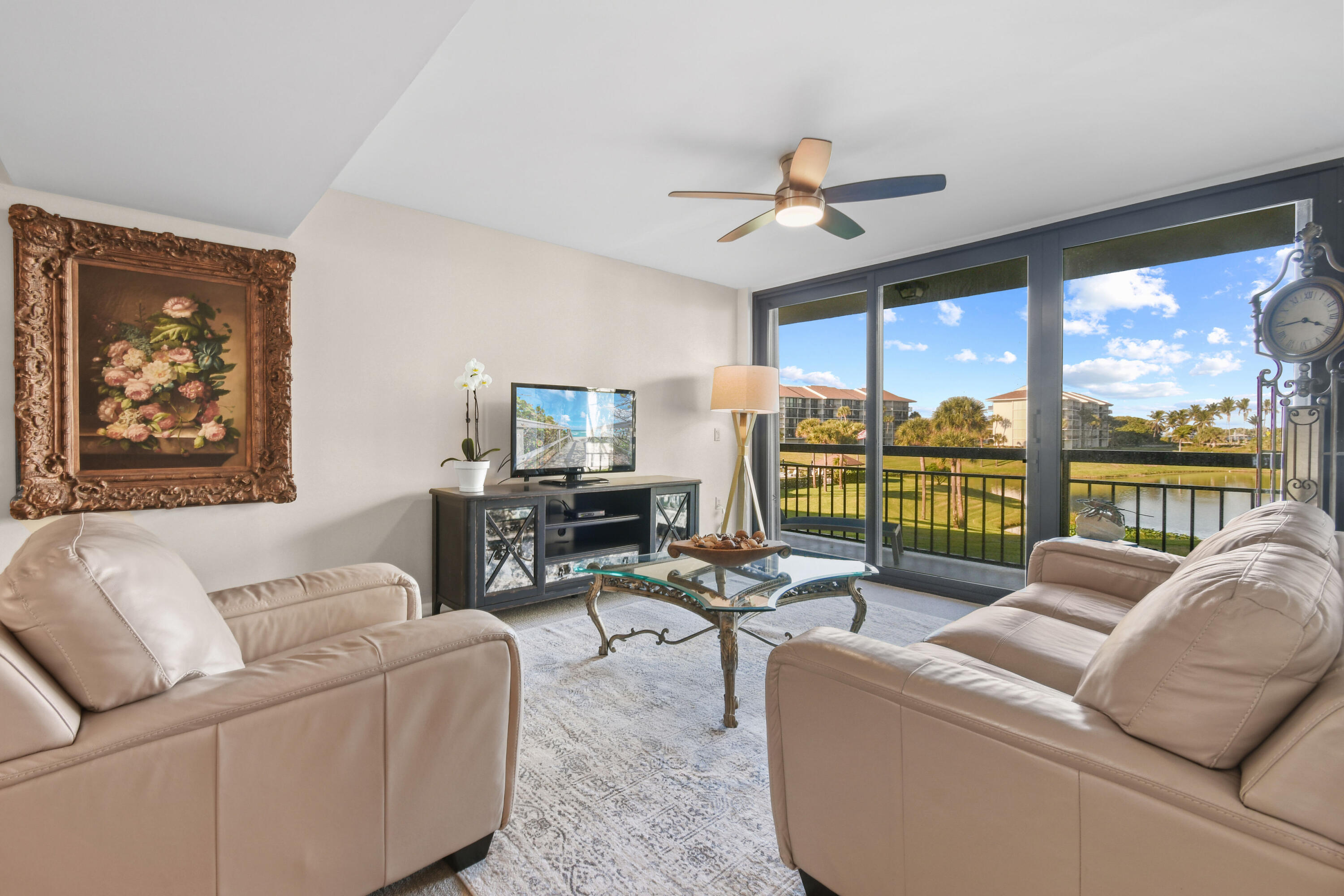 Living room with pale leather sofas and glass doors overlooking a pond and palm-lined grounds.