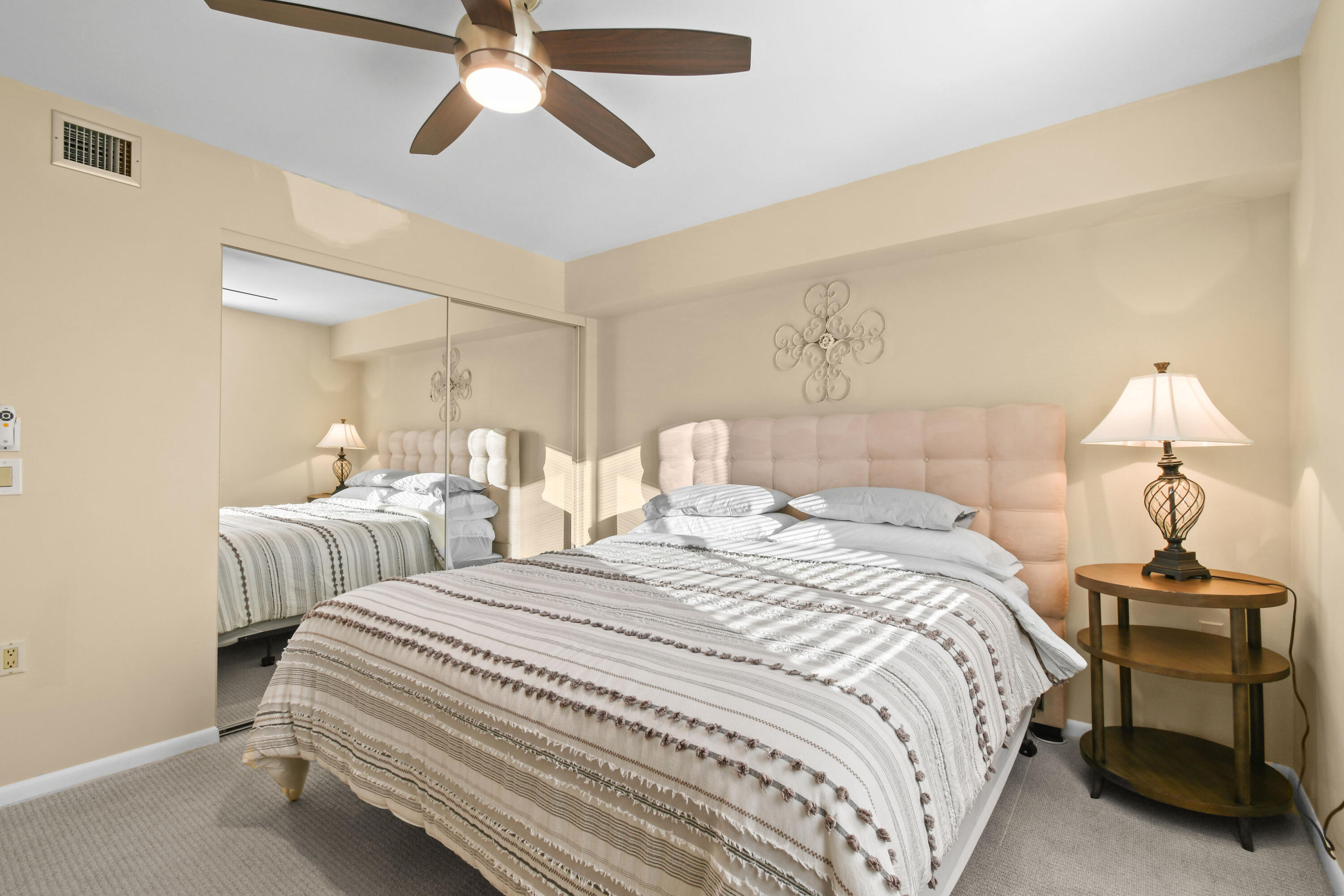 Furnished bedroom with a striped bedspread, blush tufted headboard, mirrored closet doors, and ceiling fan.