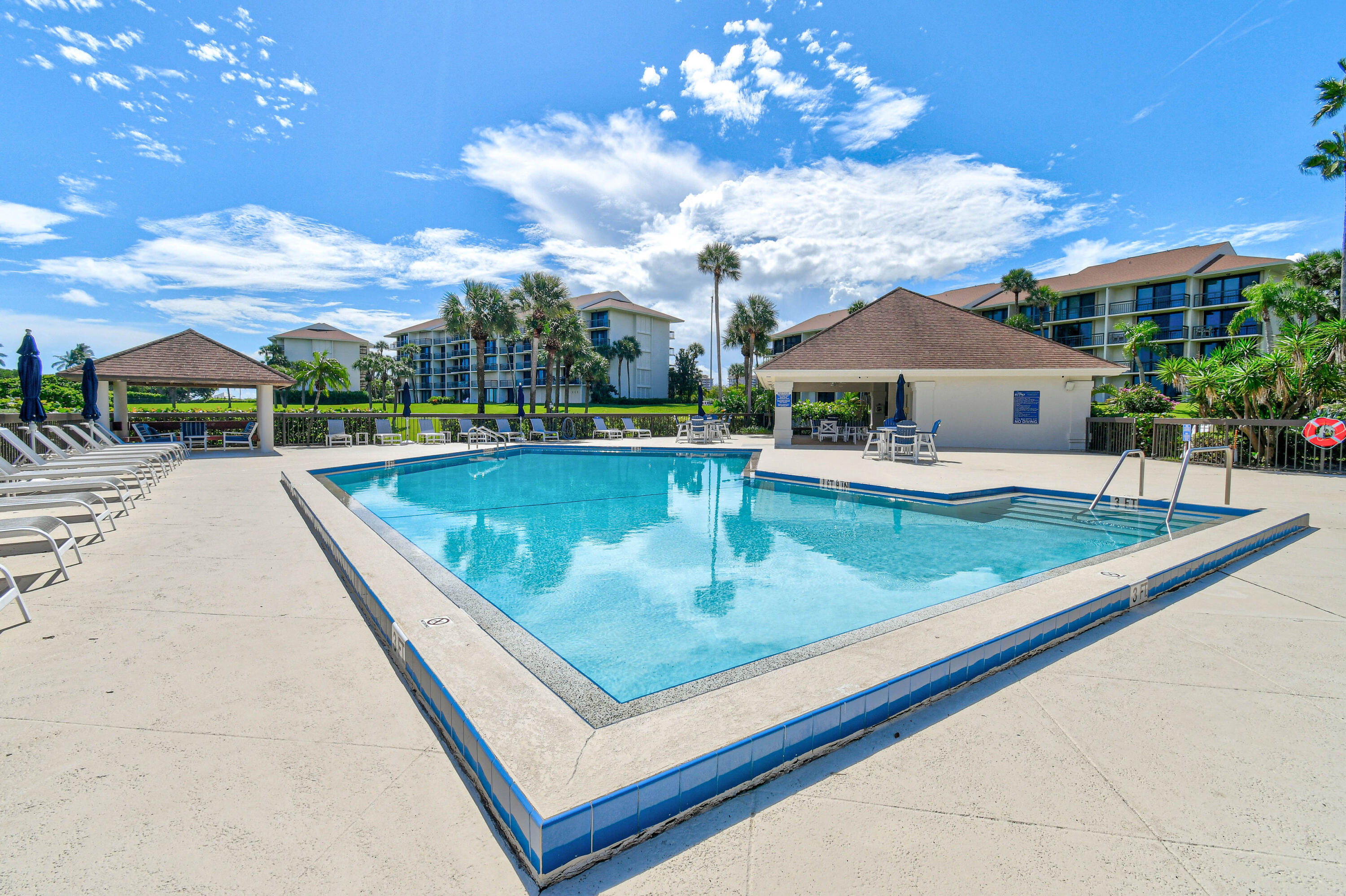 Condominium pool with blue-tiled edges, lounge chairs, shade pavilions, and palm trees.