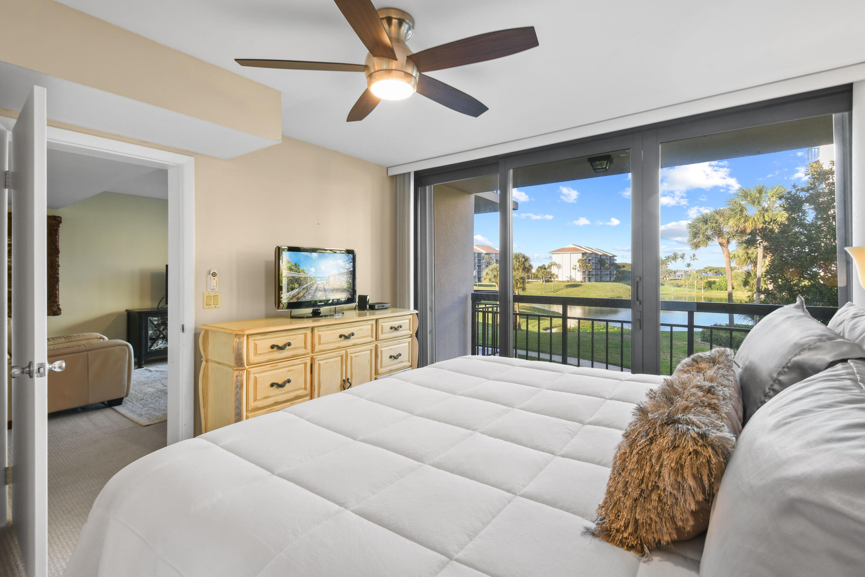 Bedroom with a white bed and sliding doors overlooking a pond and palm-lined lawn.