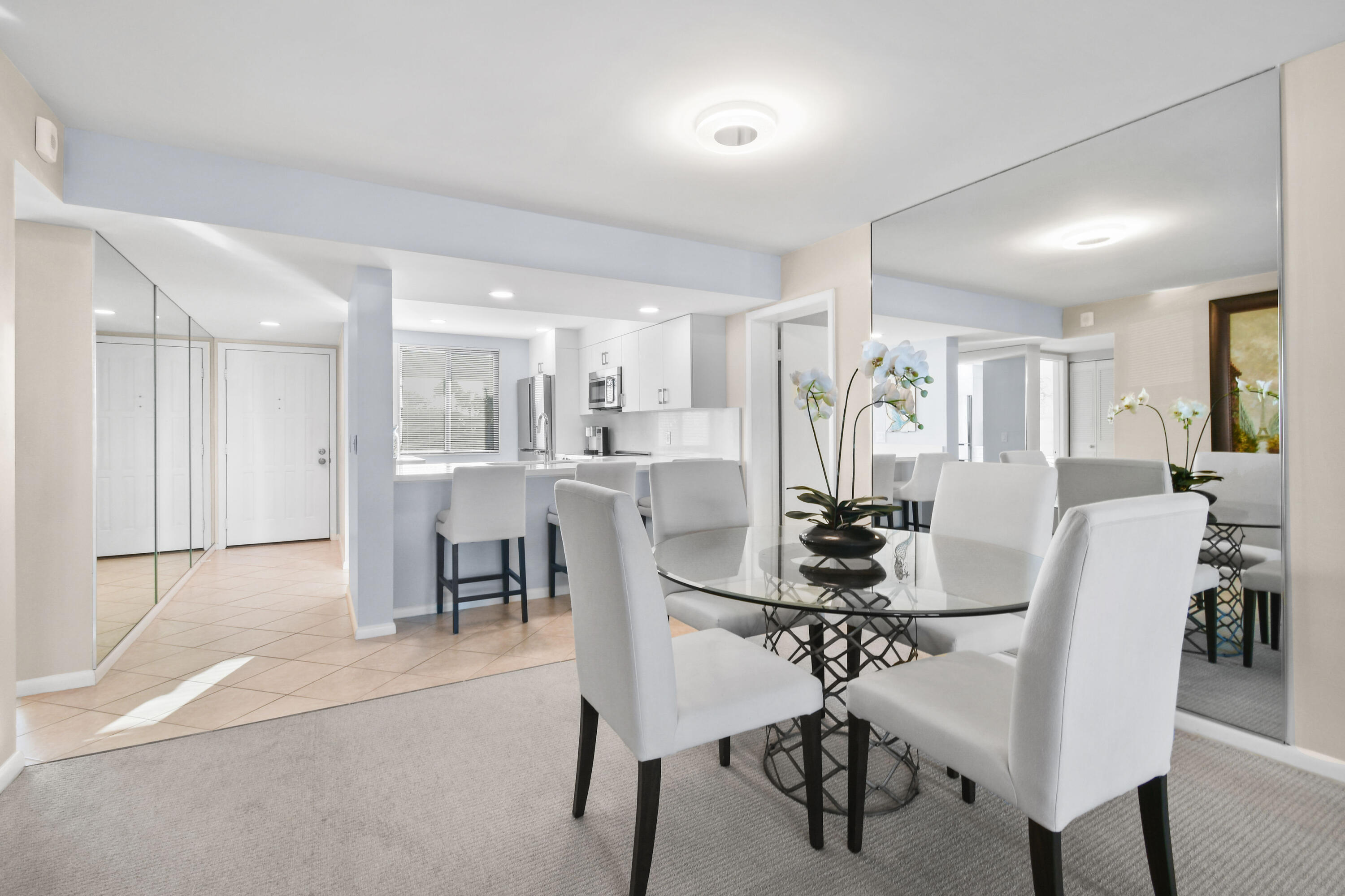 Open-plan dining area with a glass table, white chairs, mirrored walls, and an adjoining kitchen.