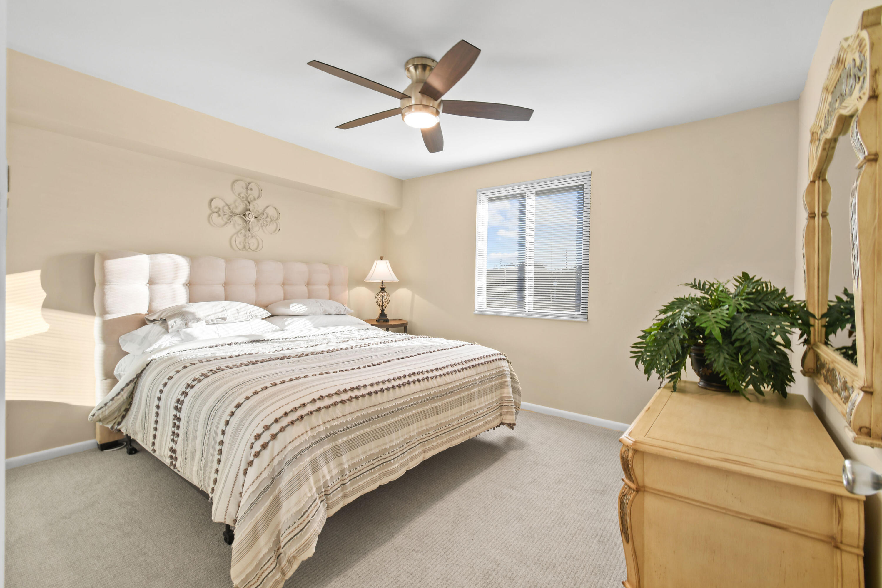 Furnished bedroom with a large tufted-headboard bed, patterned white bedding, and a dresser with a potted plant.