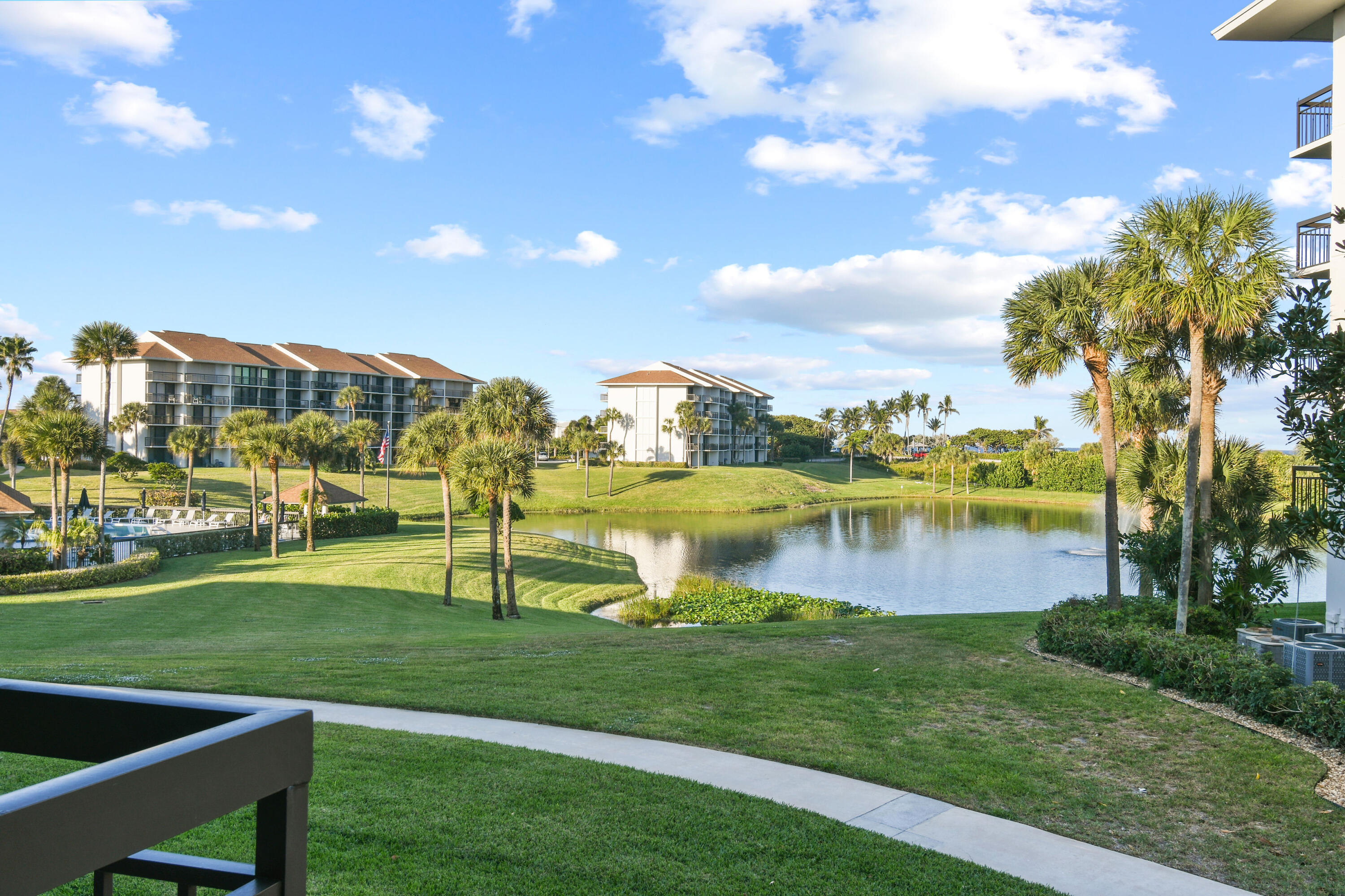 Condominium grounds with a pond, palm trees, and neighboring buildings viewed across a green lawn.