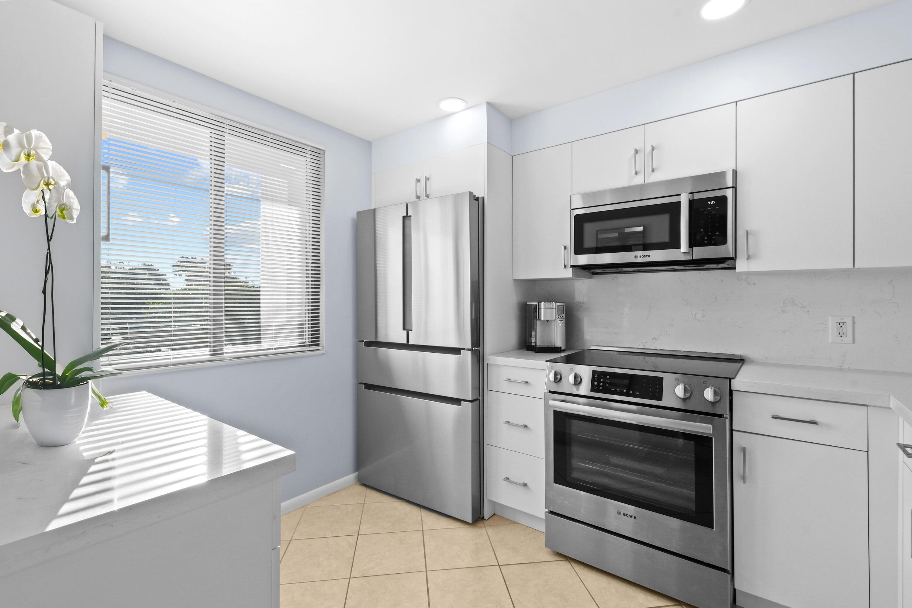 Modern white kitchen with stainless steel appliances and an orchid beside a sunlit window.