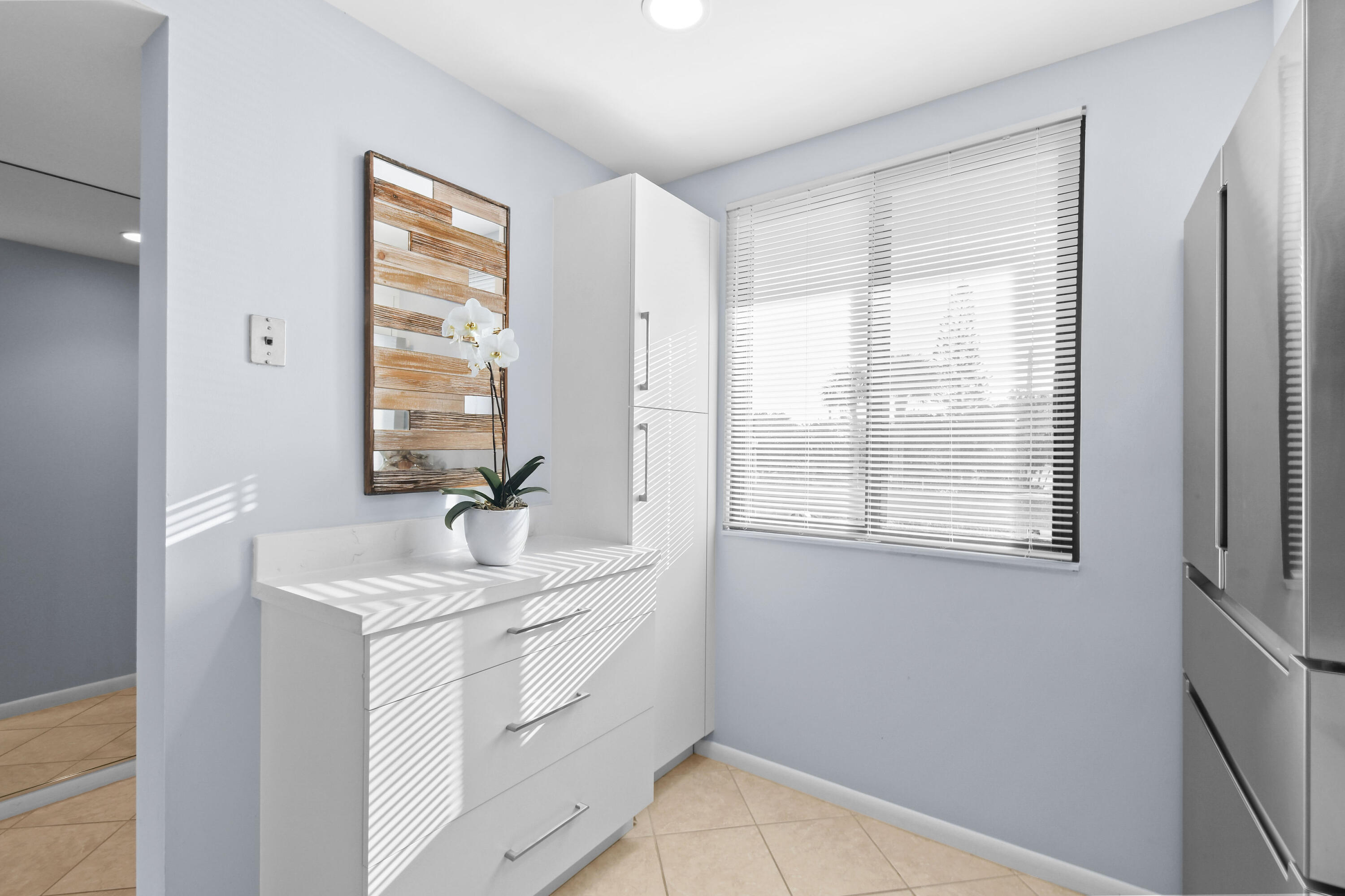 Bright kitchen with white cabinetry, stainless steel refrigerator, and sunlight through window blinds.