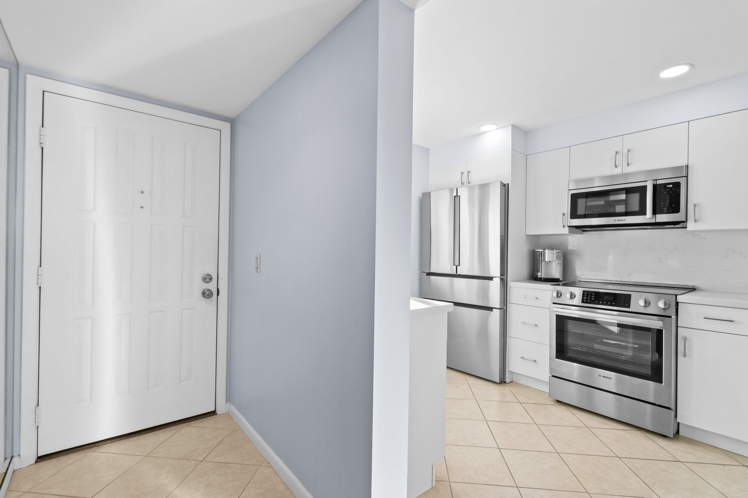 Condo entryway beside a modern white kitchen with stainless steel appliances and beige tile flooring.