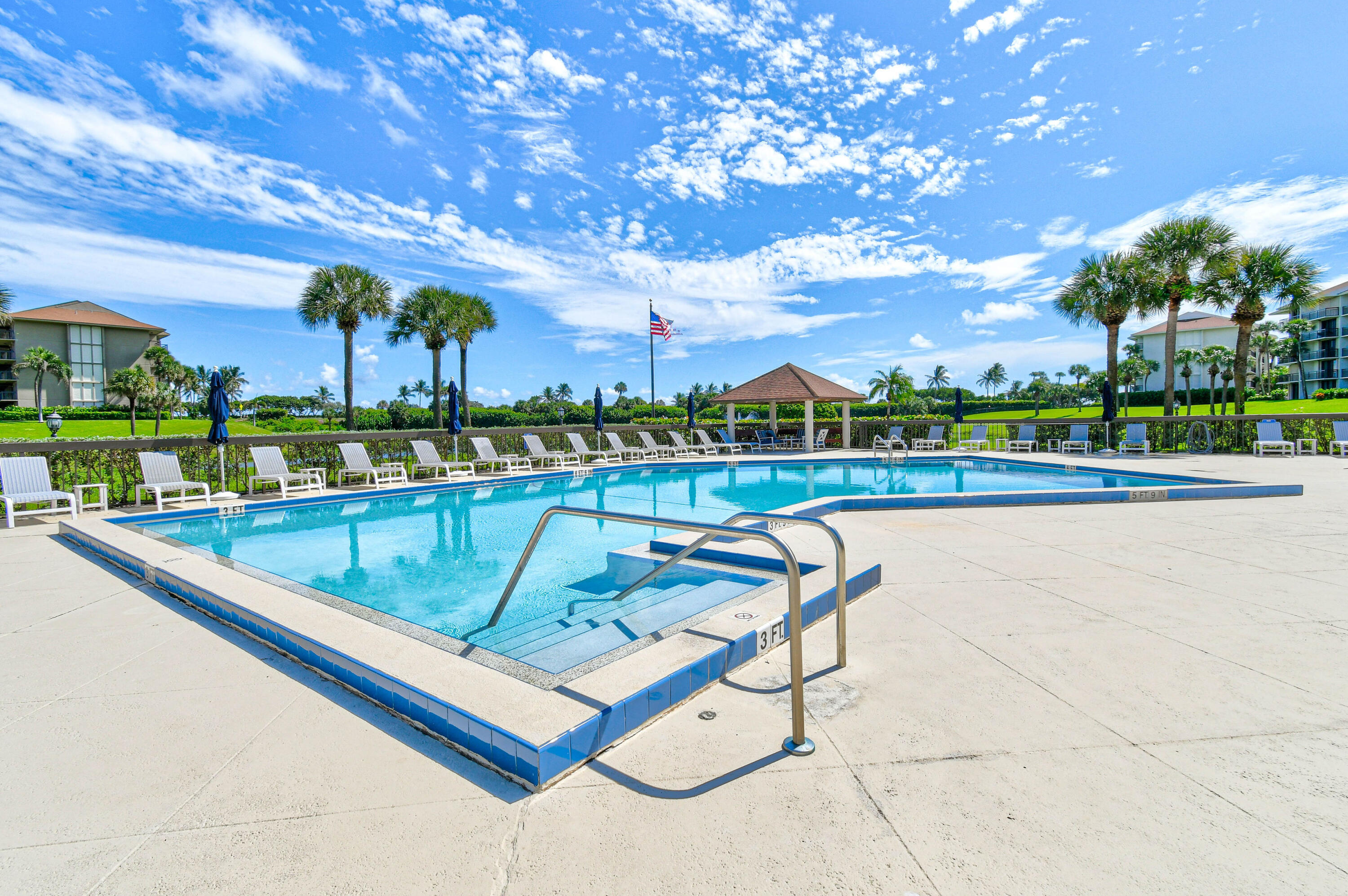 Community pool with metal entry rails, lounge chairs, a gazebo, and palm trees.