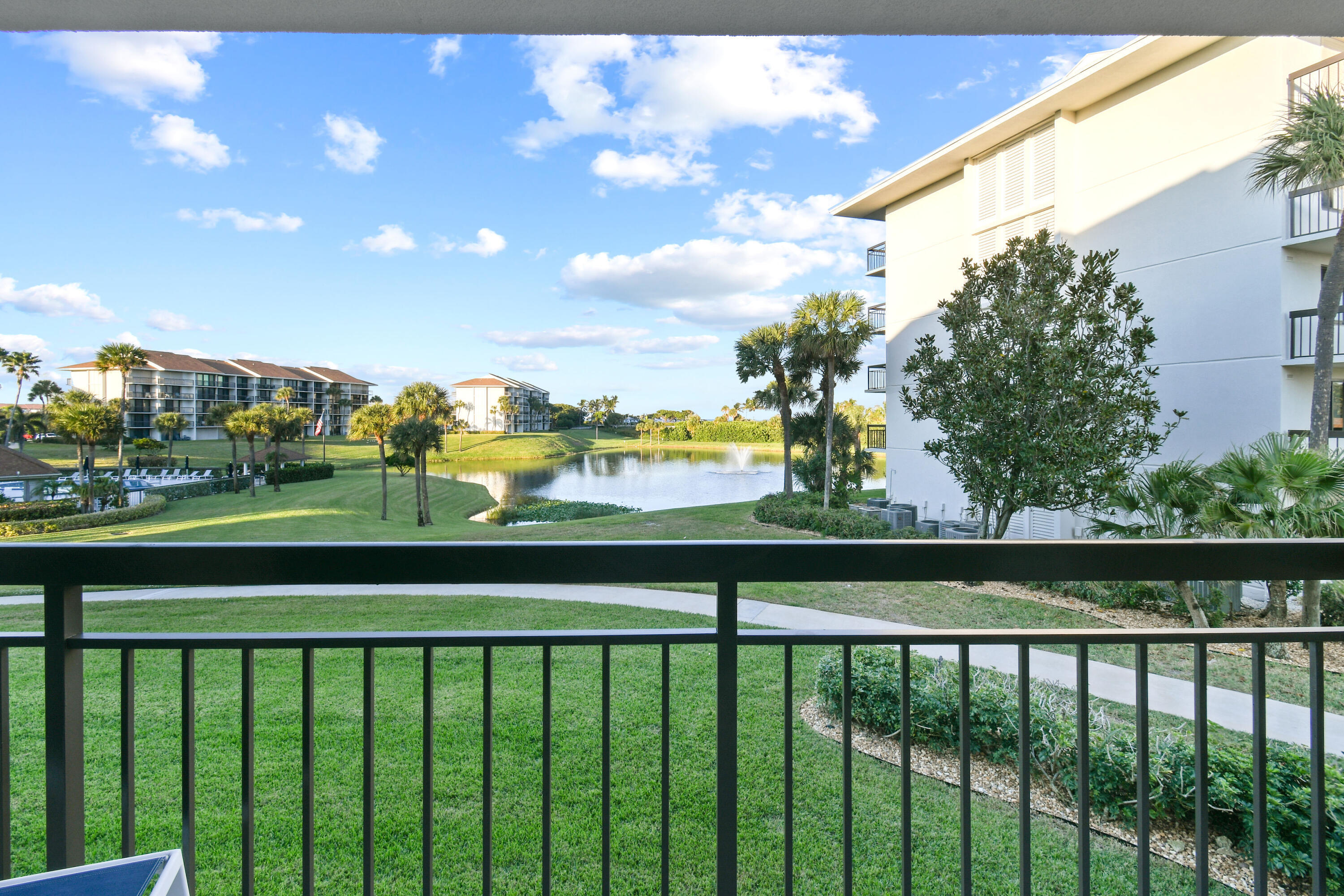 Condo balcony overlooks a pond with a fountain, landscaped lawns, palms, and neighboring buildings.