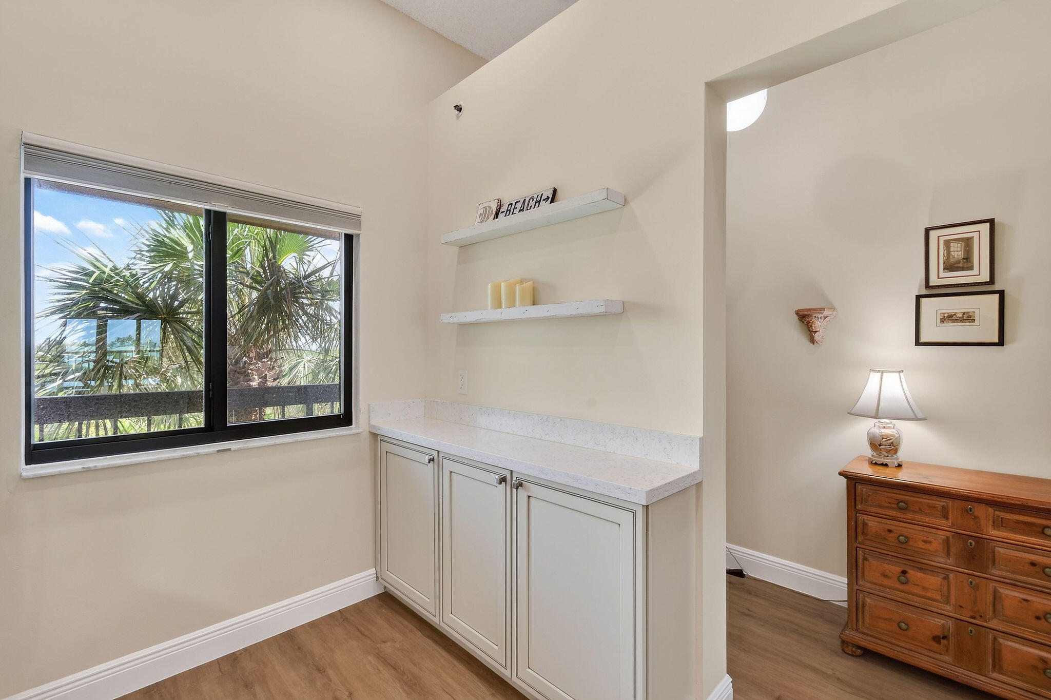 Bedroom sitting area with white built-ins beside a palm-framed window and wooden dresser.
