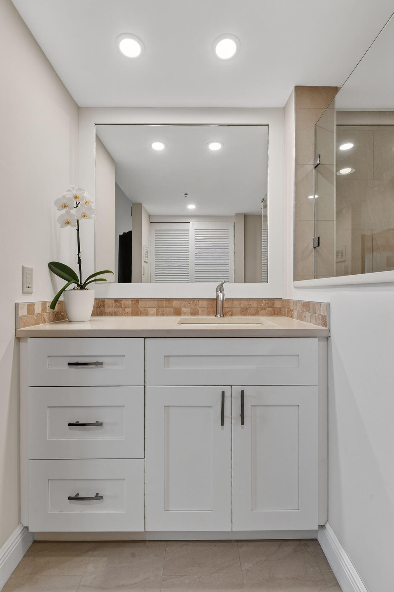 Bathroom vanity with white cabinets, tan tile backsplash, large mirror, and a white orchid beside the sink.