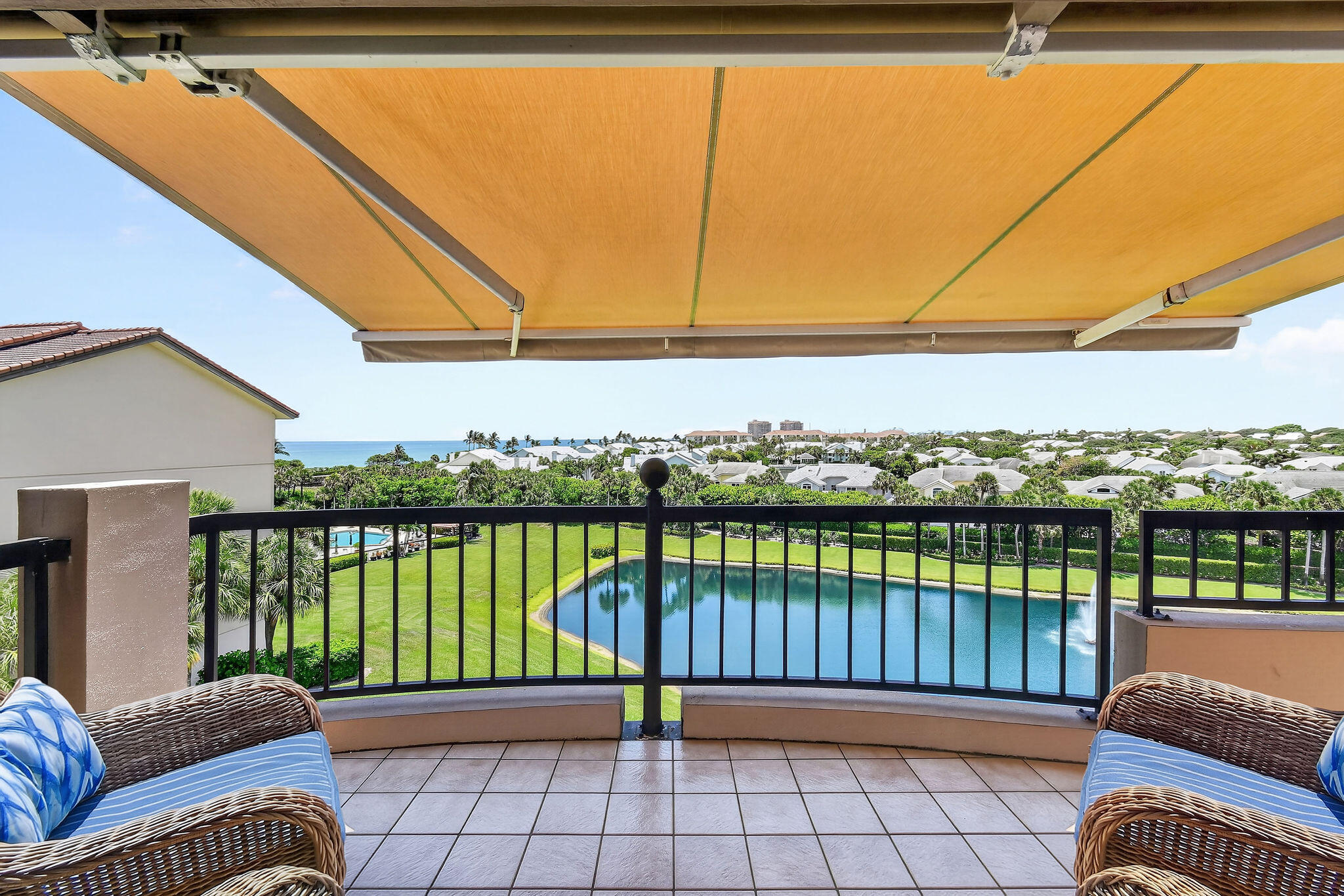 Furnished balcony with an ochre awning overlooking a pool, homes, and distant ocean.