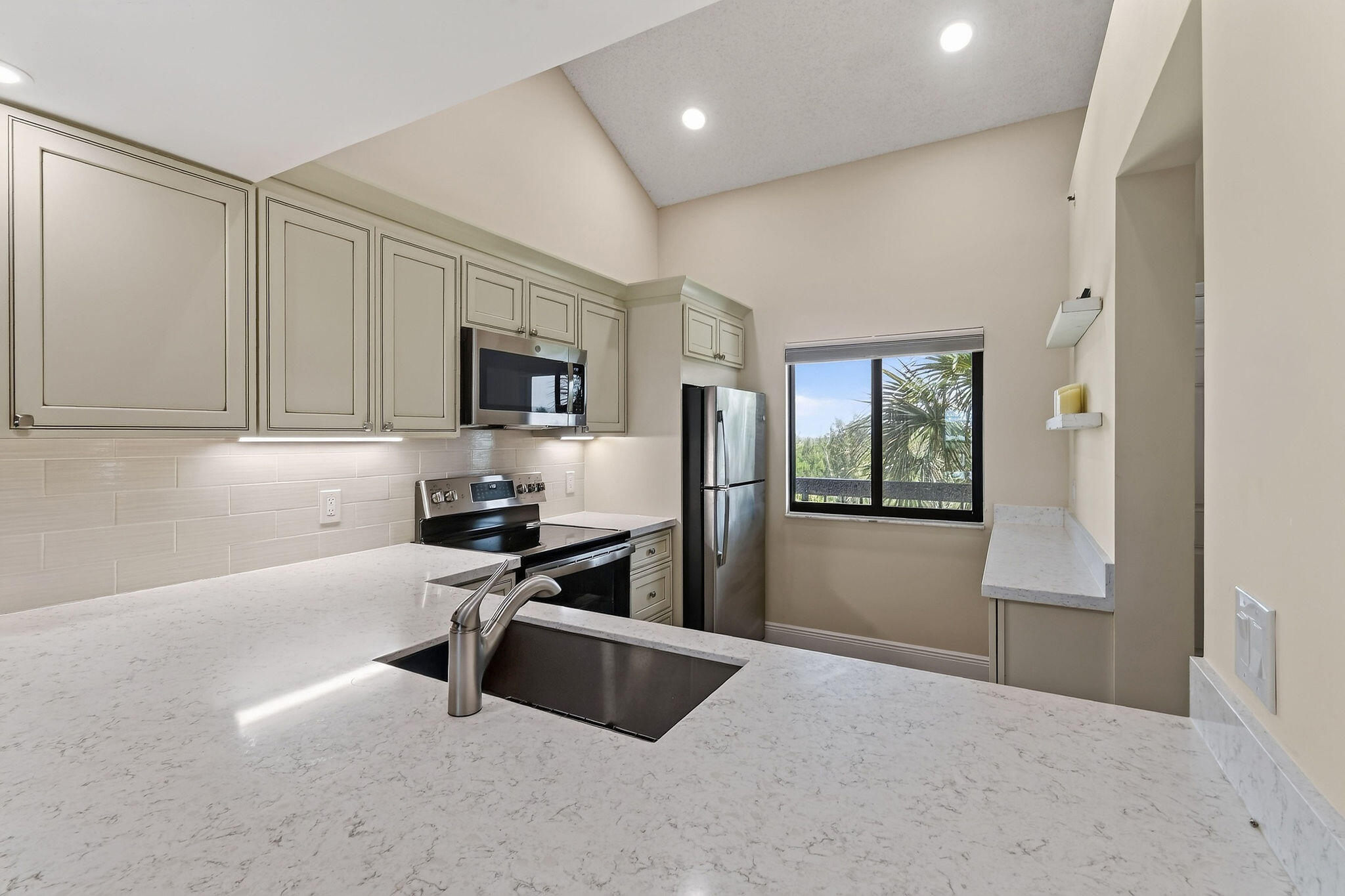 Kitchen with white stone counters, pale cabinets, stainless appliances, and a window overlooking palm trees.