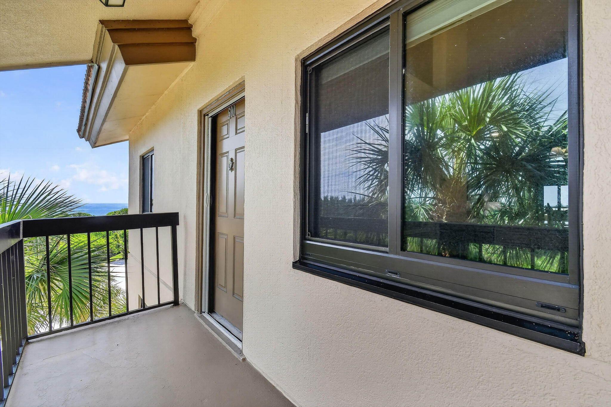 Covered condo balcony with a black railing, palm trees, and a glimpse of ocean.