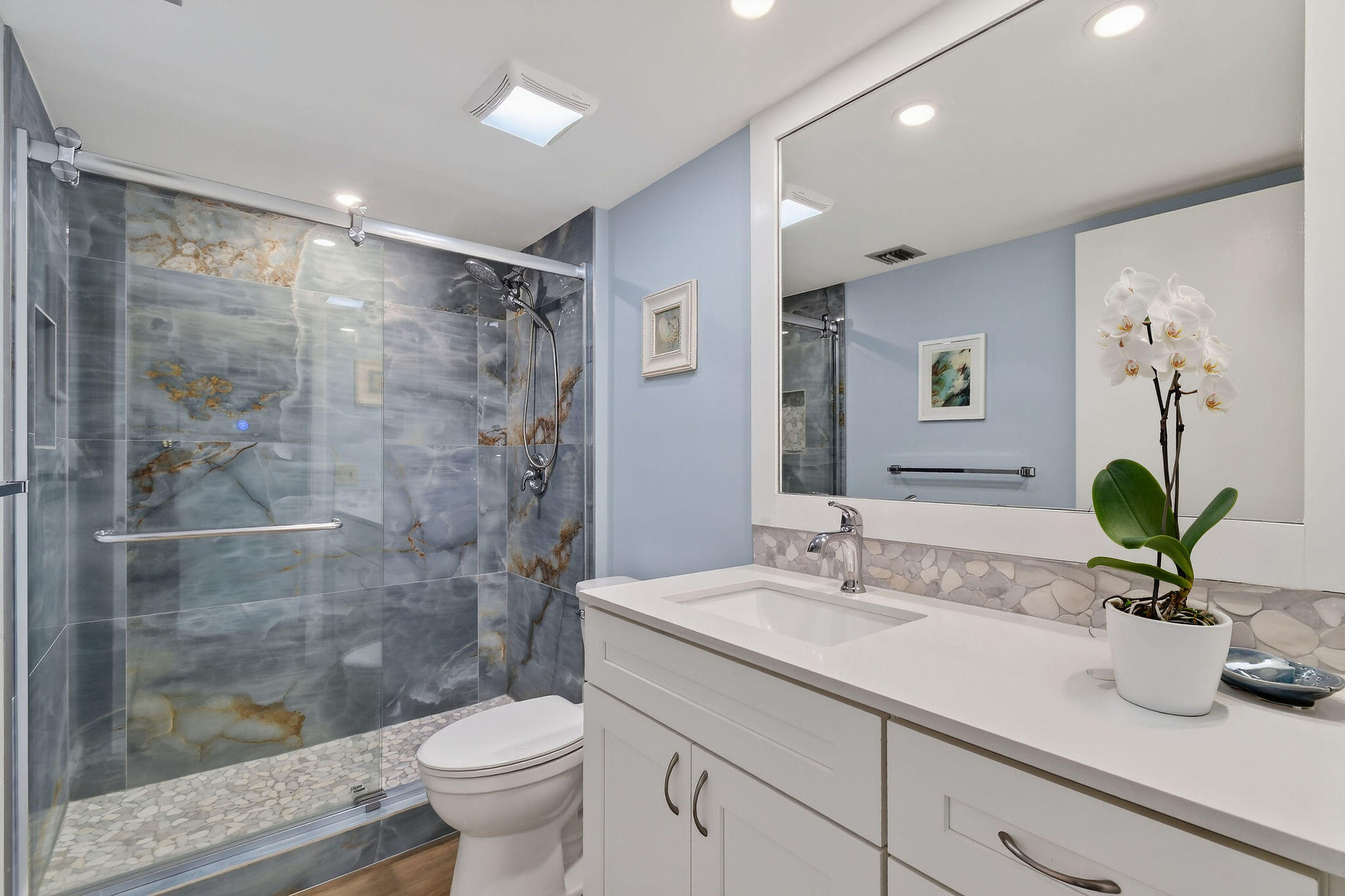 Bathroom with a veined-tile walk-in shower, white vanity, and potted orchid.