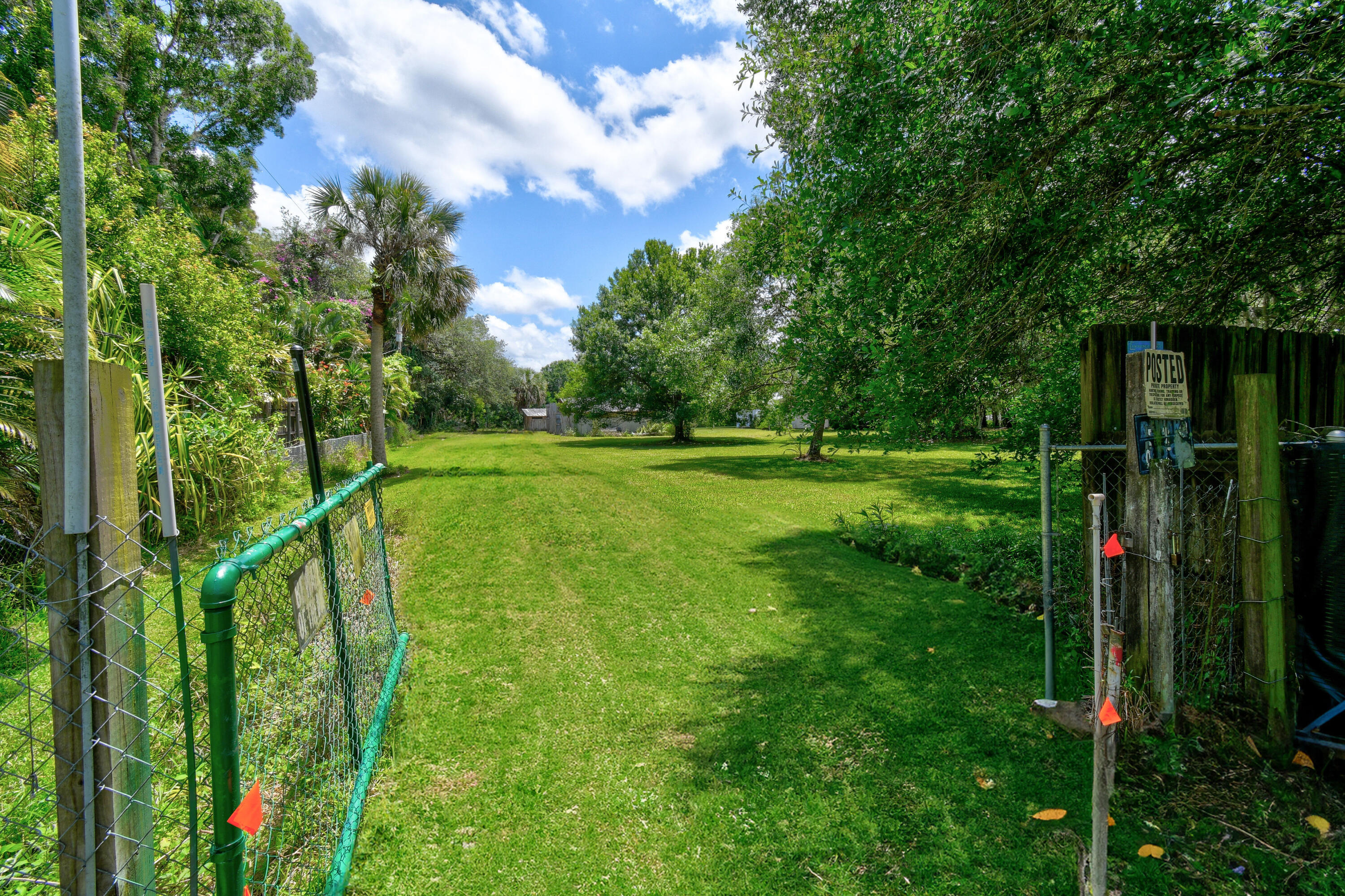 Grassy property lot bordered by chain-link fences, tropical landscaping, and mature trees.