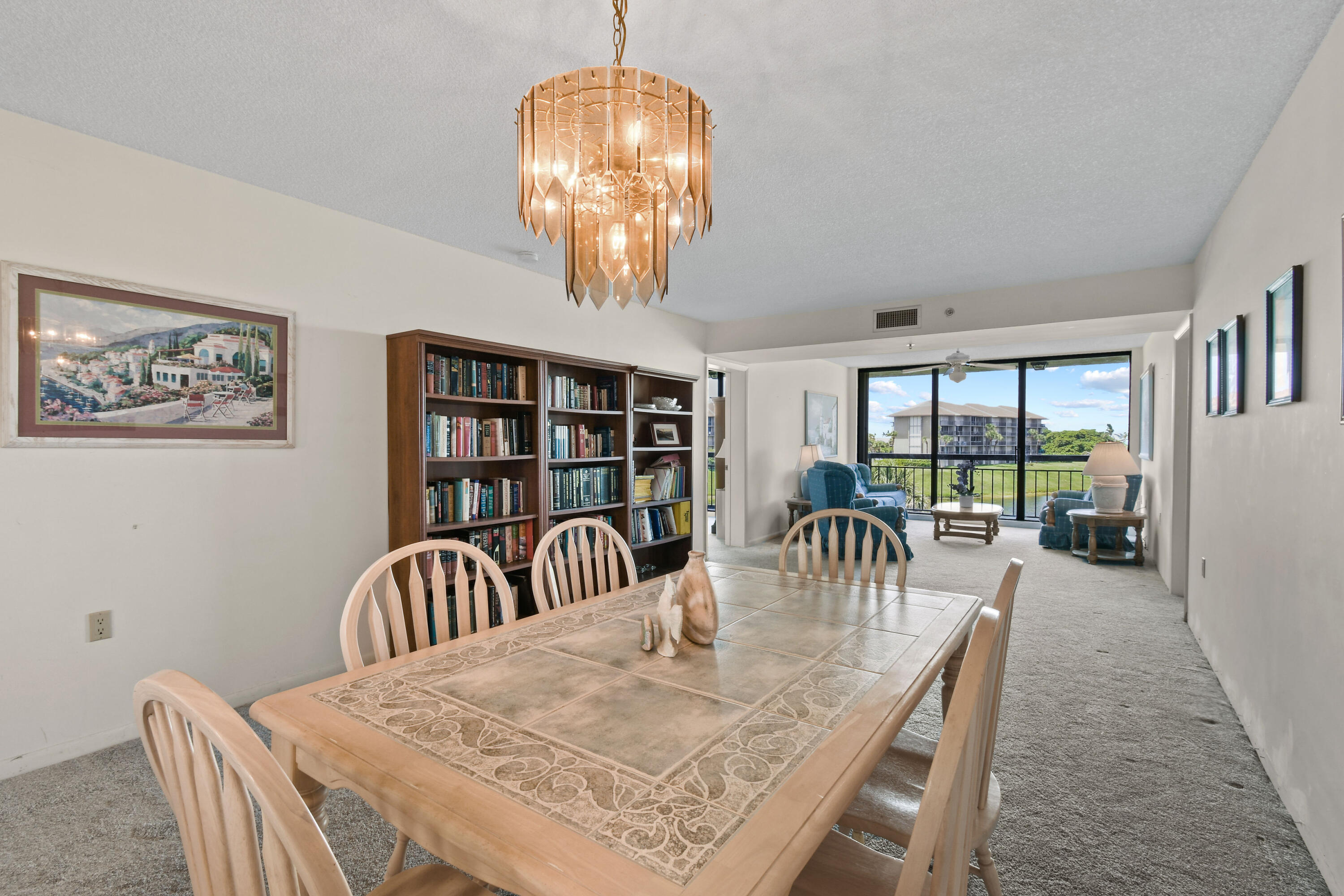 Condo dining area with a tiled table and chandelier, opening to a living room with balcony views.