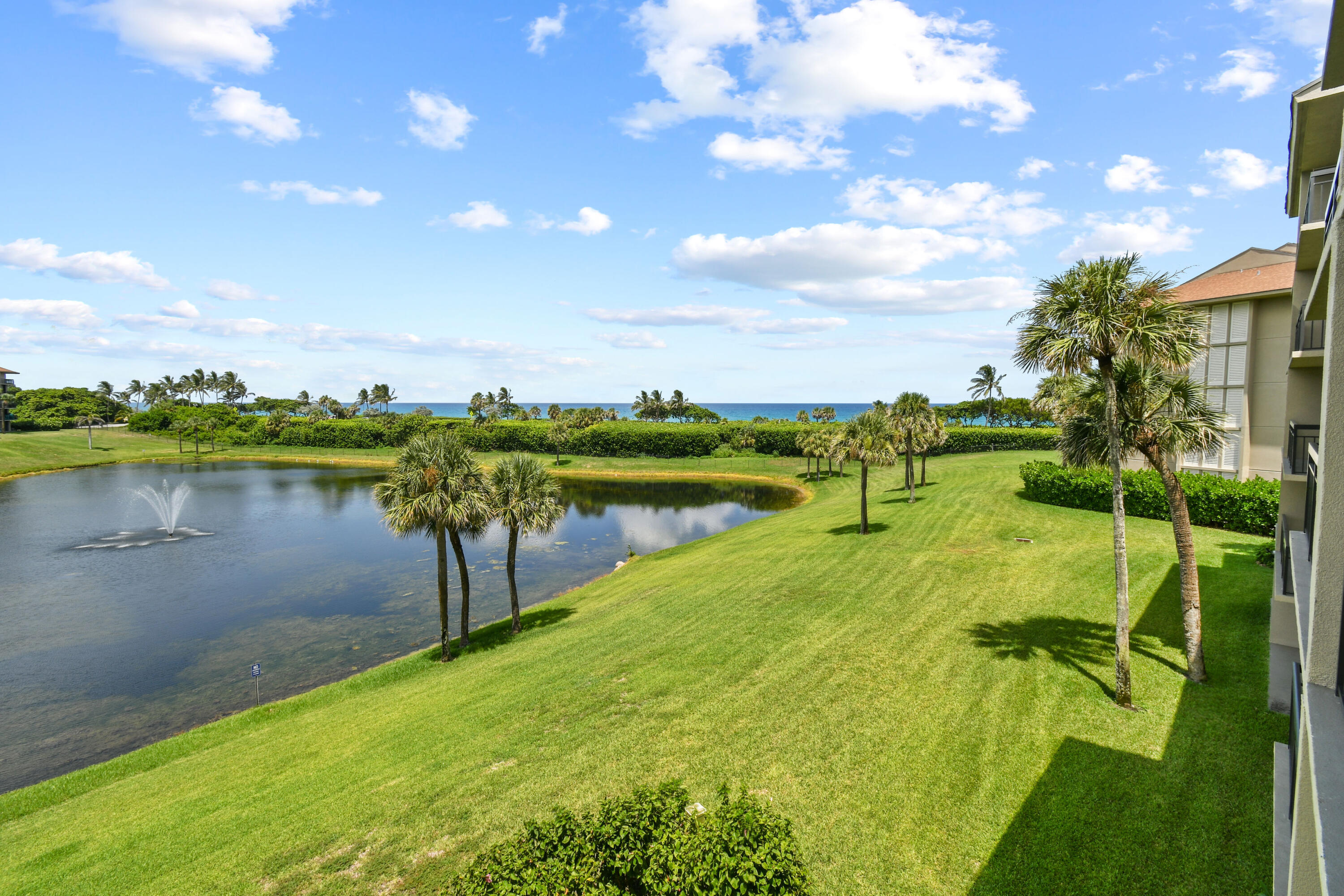 Landscaped pond and lawn with palm trees, a fountain, and ocean views beside a condominium.