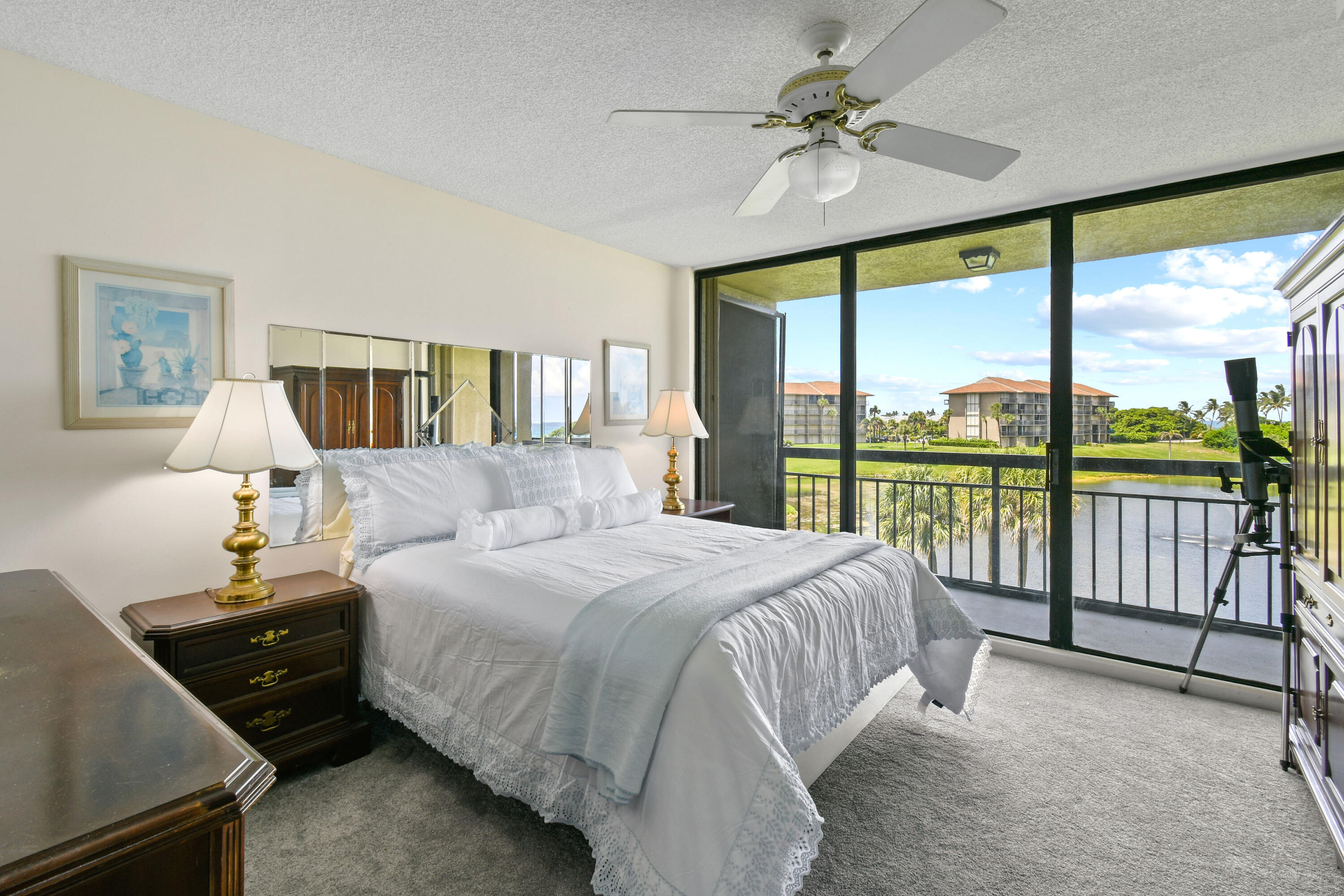 Bedroom with a white bed and sliding glass doors opening to a balcony overlooking a pond and landscaped grounds.