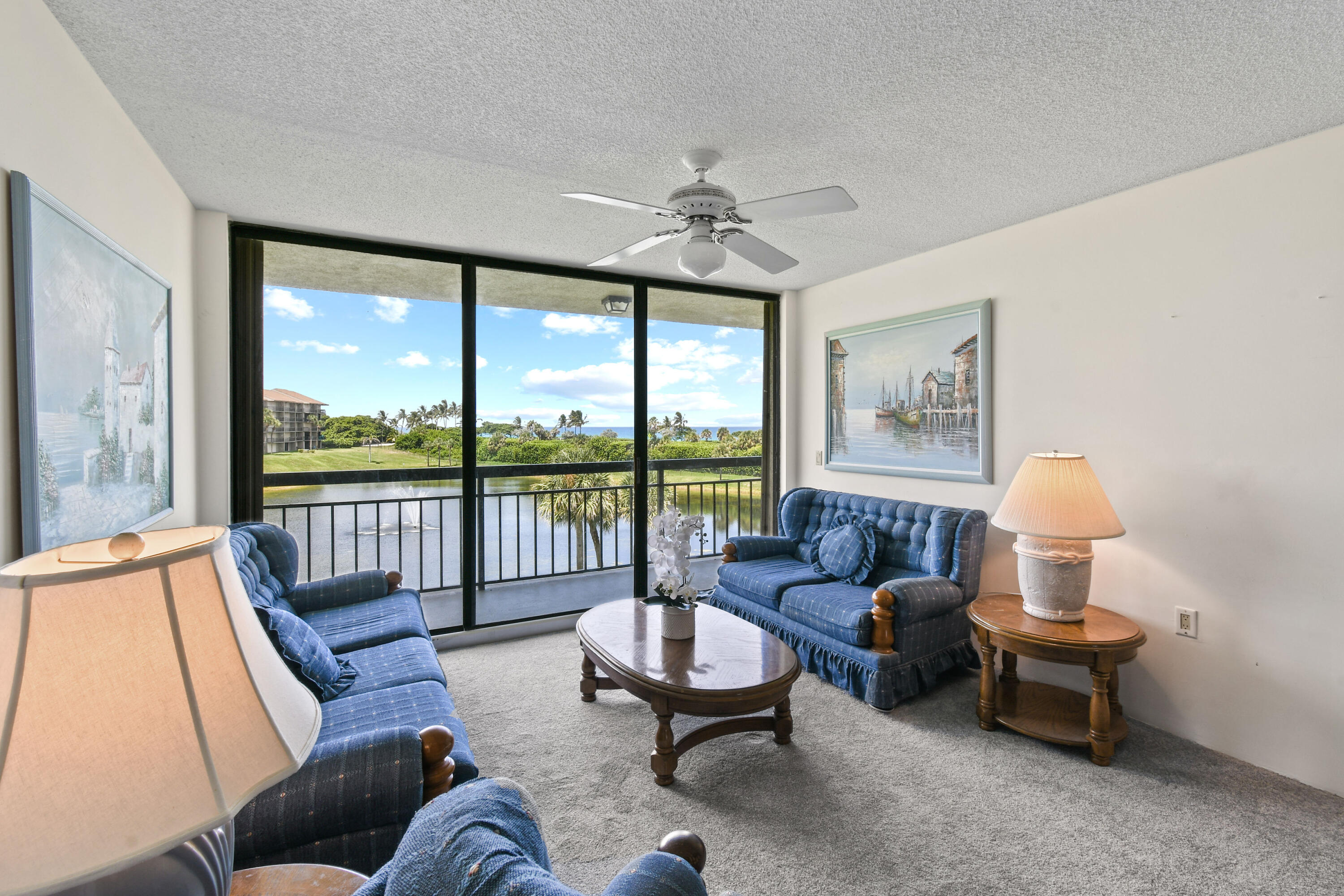 Living room with blue sofas and balcony doors overlooking a waterway, palm trees, and distant ocean.