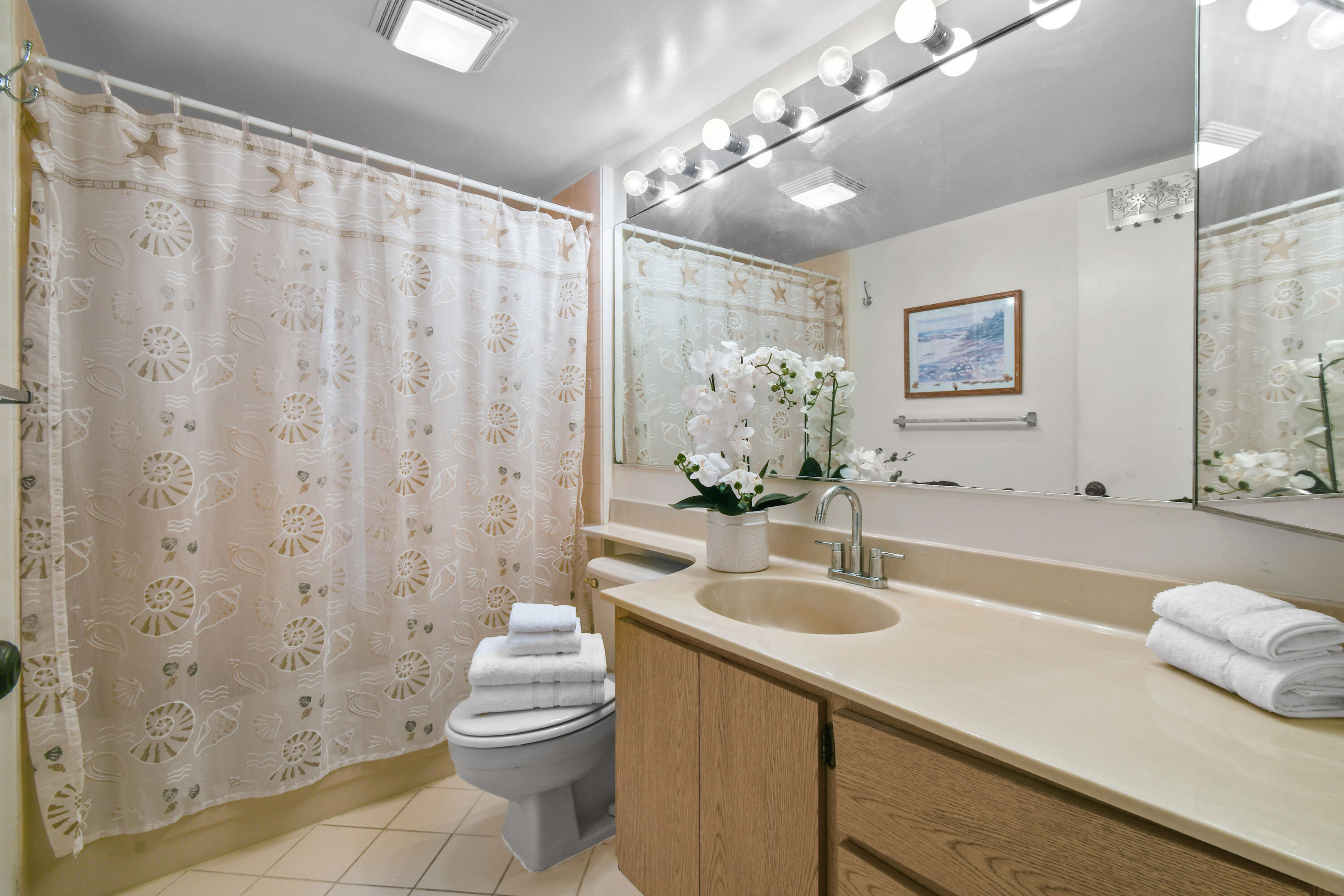 Bathroom with a seashell-patterned shower curtain, long vanity, large mirror, and white orchids.