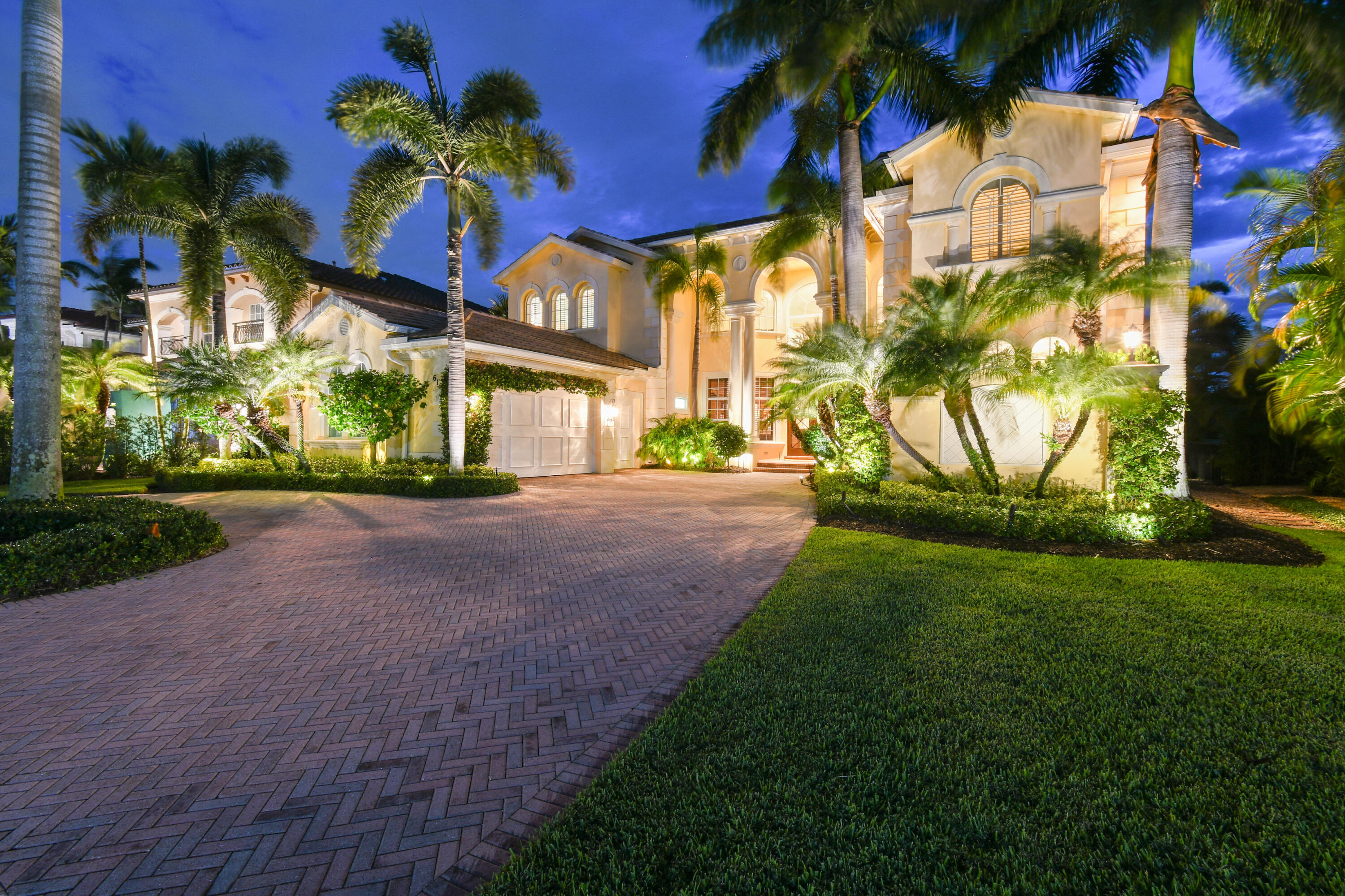 Two-story stucco home with arched windows, a broad brick driveway, and palm trees at dusk.