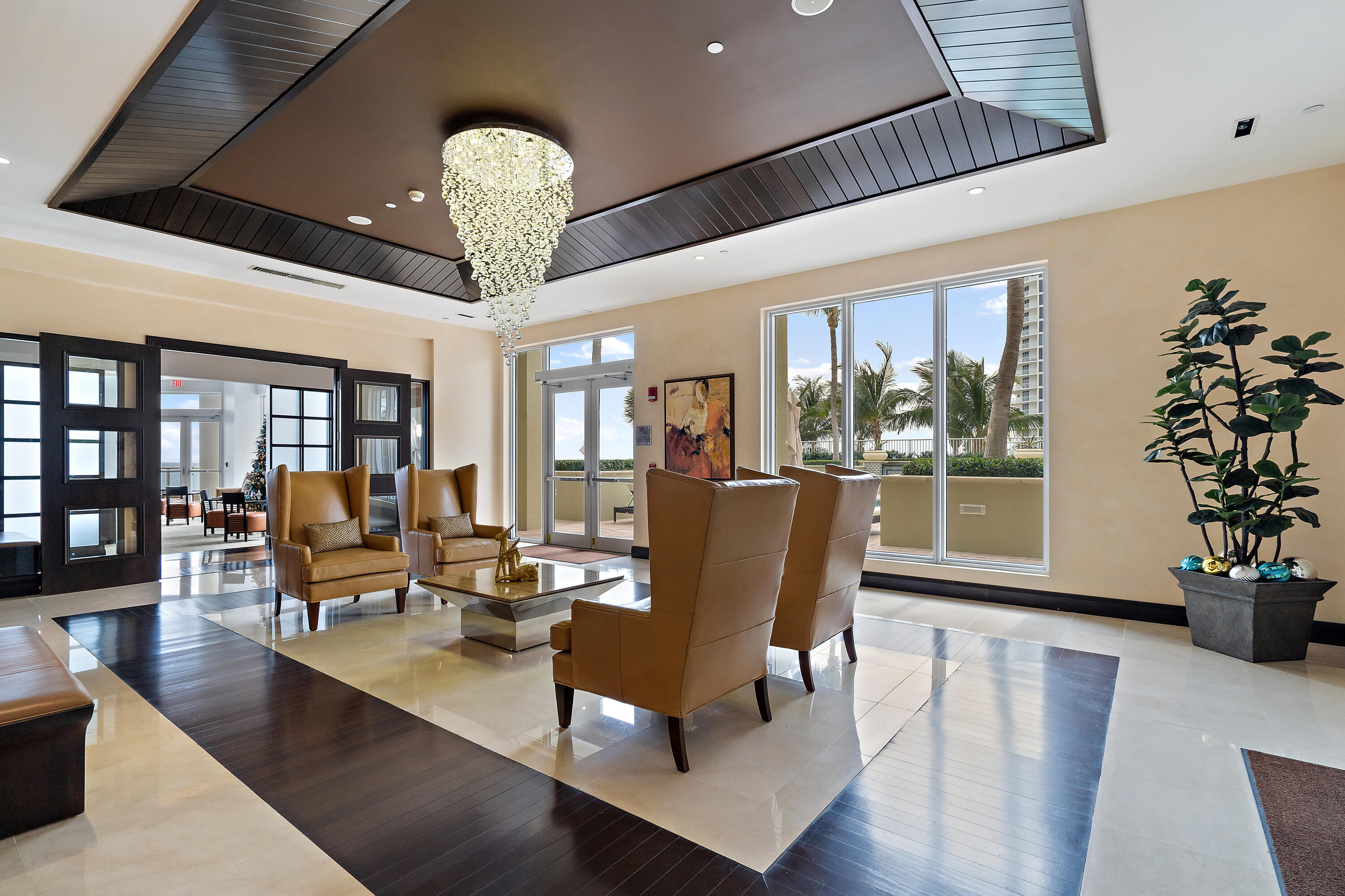Condominium lobby lounge with tan leather chairs, a crystal chandelier, and palm trees outside the windows.