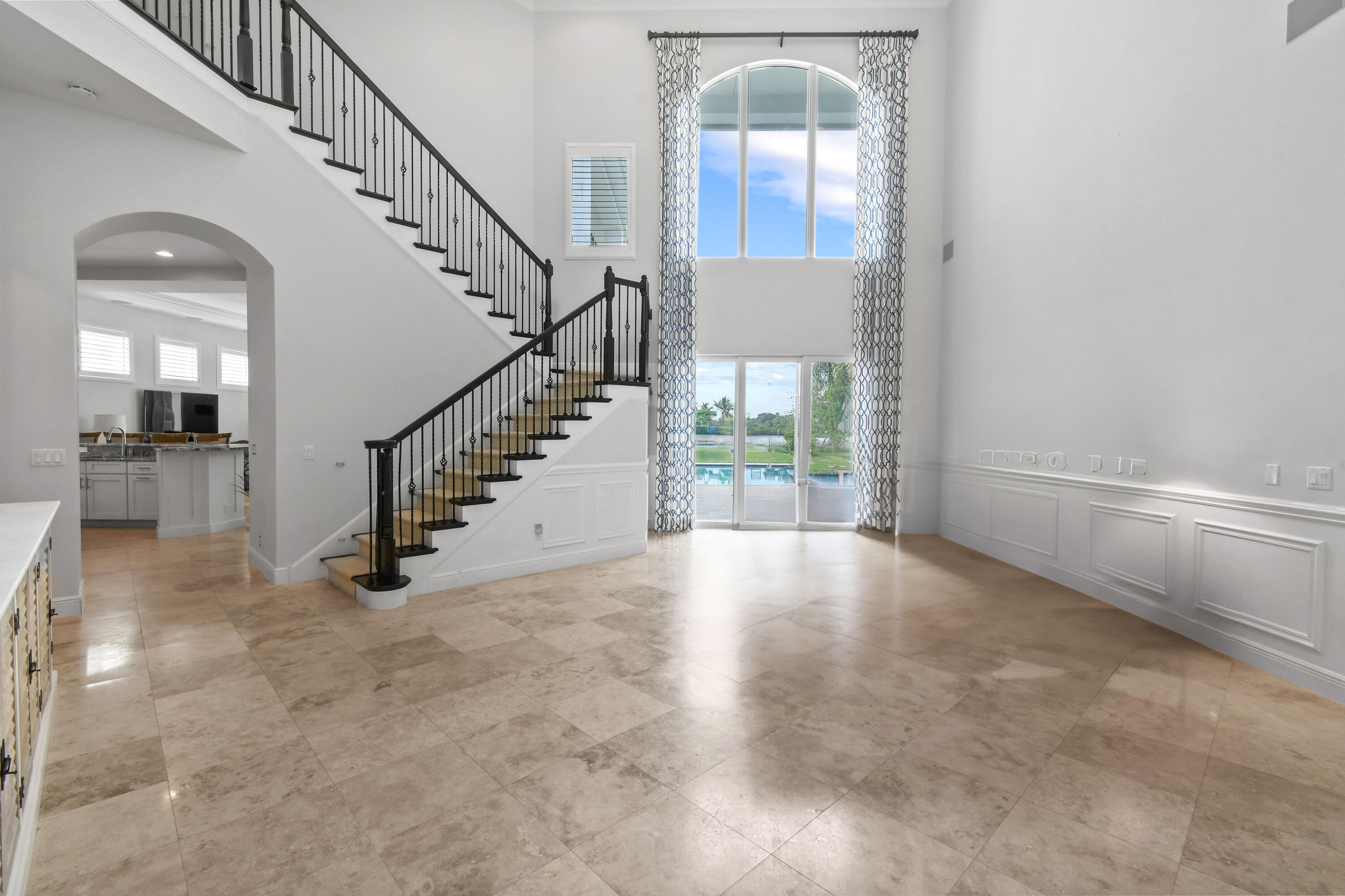 Two-story living area with a sweeping staircase, stone tile floors, and tall windows overlooking a pool.