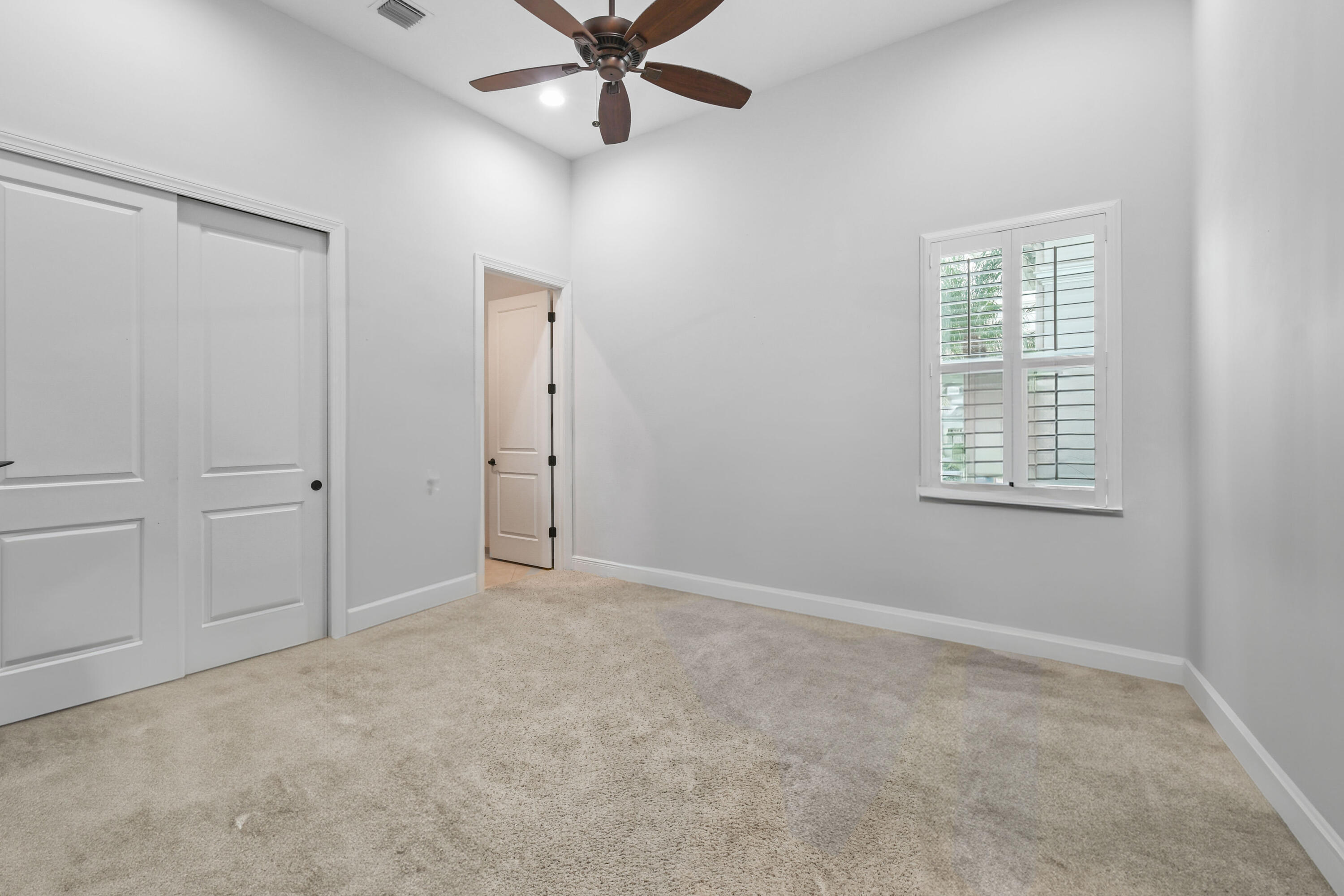 Empty carpeted bedroom with a ceiling fan, sliding closet doors, and a shuttered window.