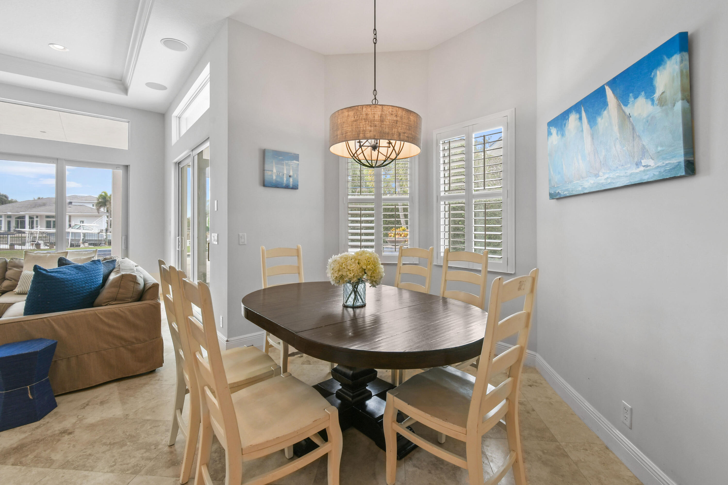 Dining area with an oval table, light wood chairs, shuttered windows, and a drum chandelier beside the living room.