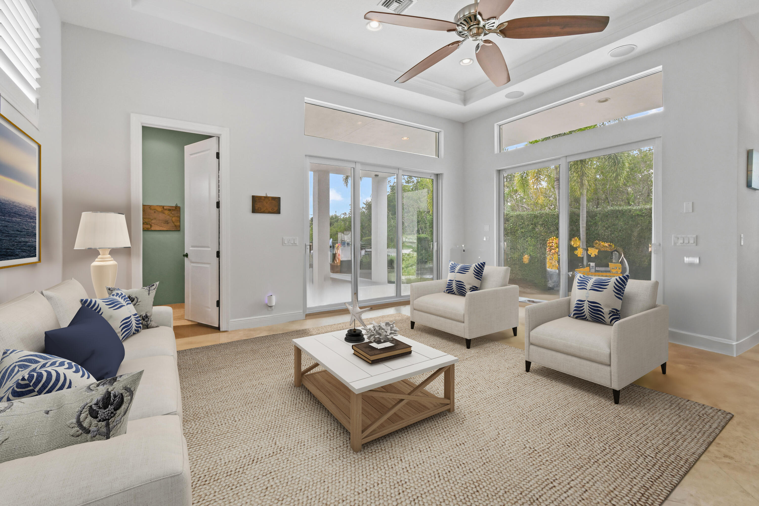 Staged living room with blue-and-white seating, a woven rug, and large windows overlooking tropical landscaping.