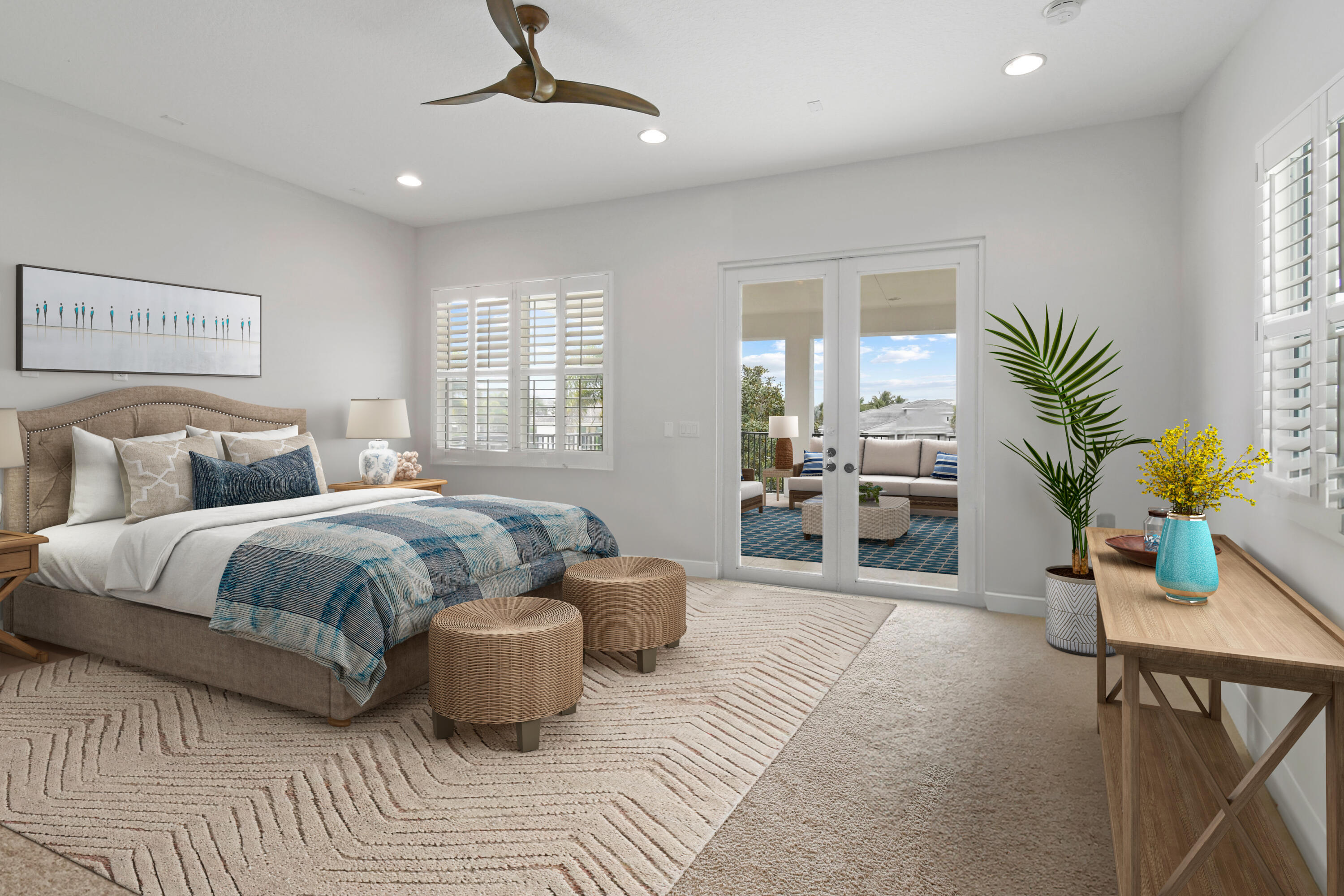 Staged bedroom with blue bedding, woven ottomans, and French doors to a furnished balcony.
