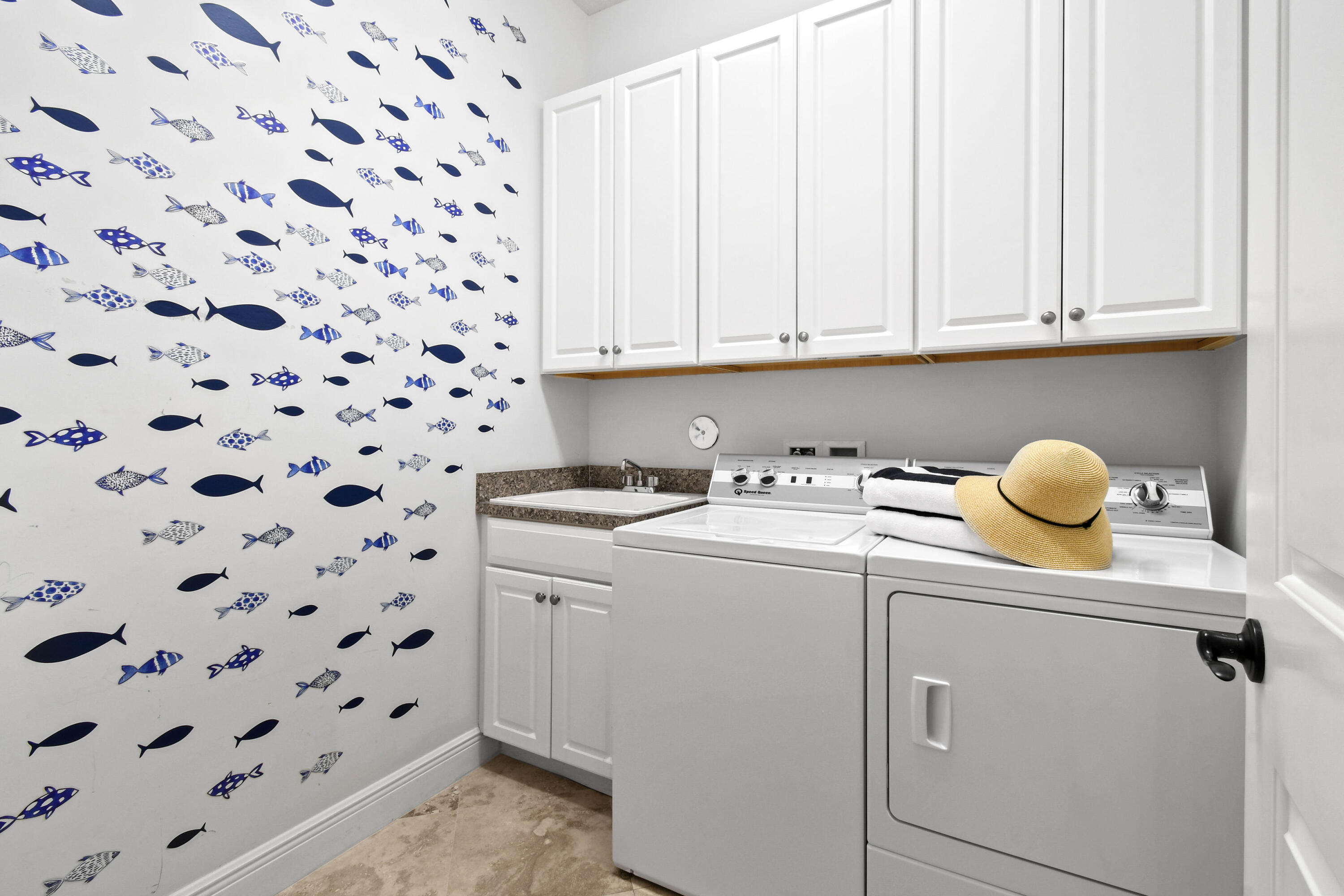 Laundry room with fish-patterned wall, white washer and dryer, and a straw hat atop folded towels.