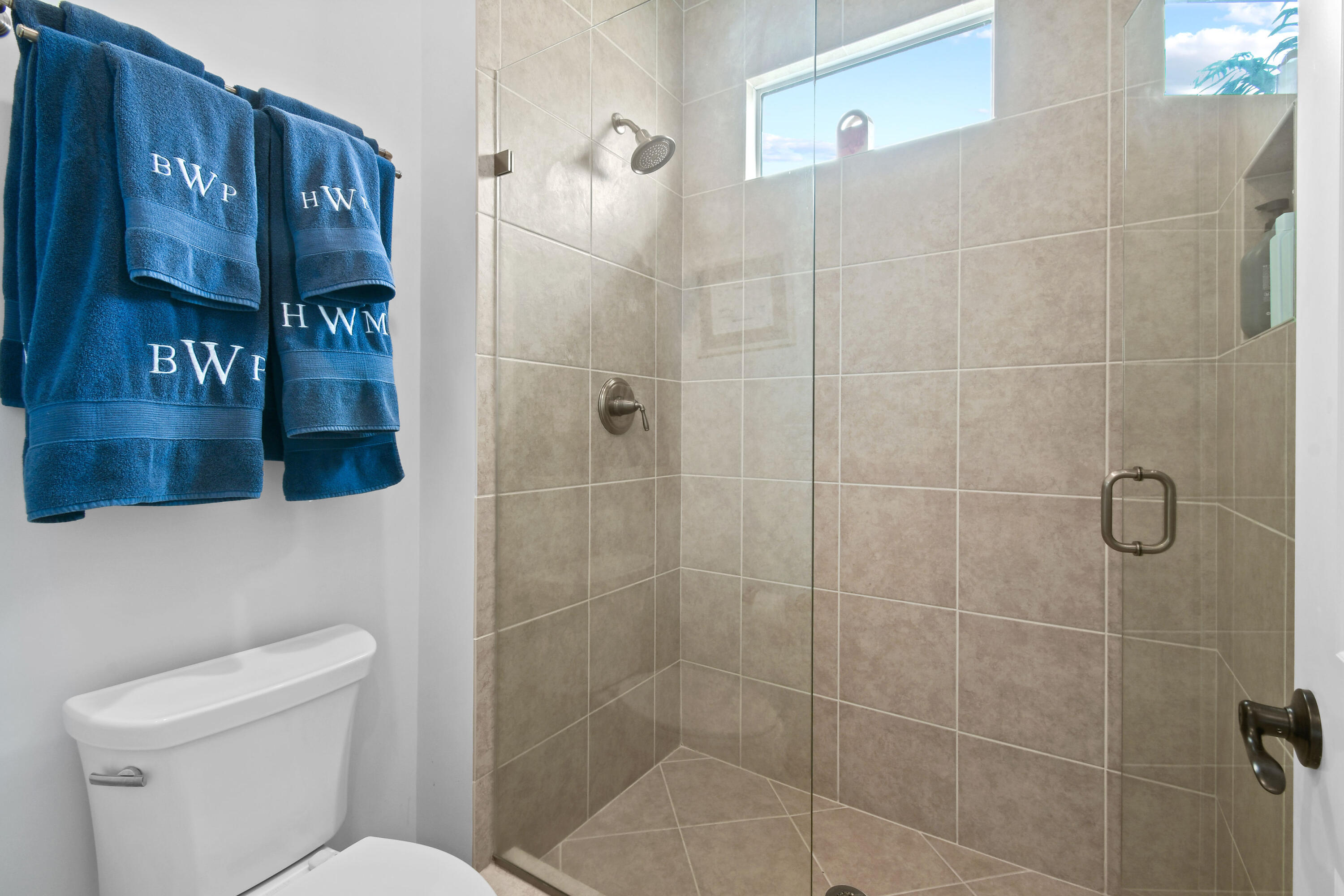 Tiled walk-in shower with a high window and blue monogrammed towels above a white toilet.