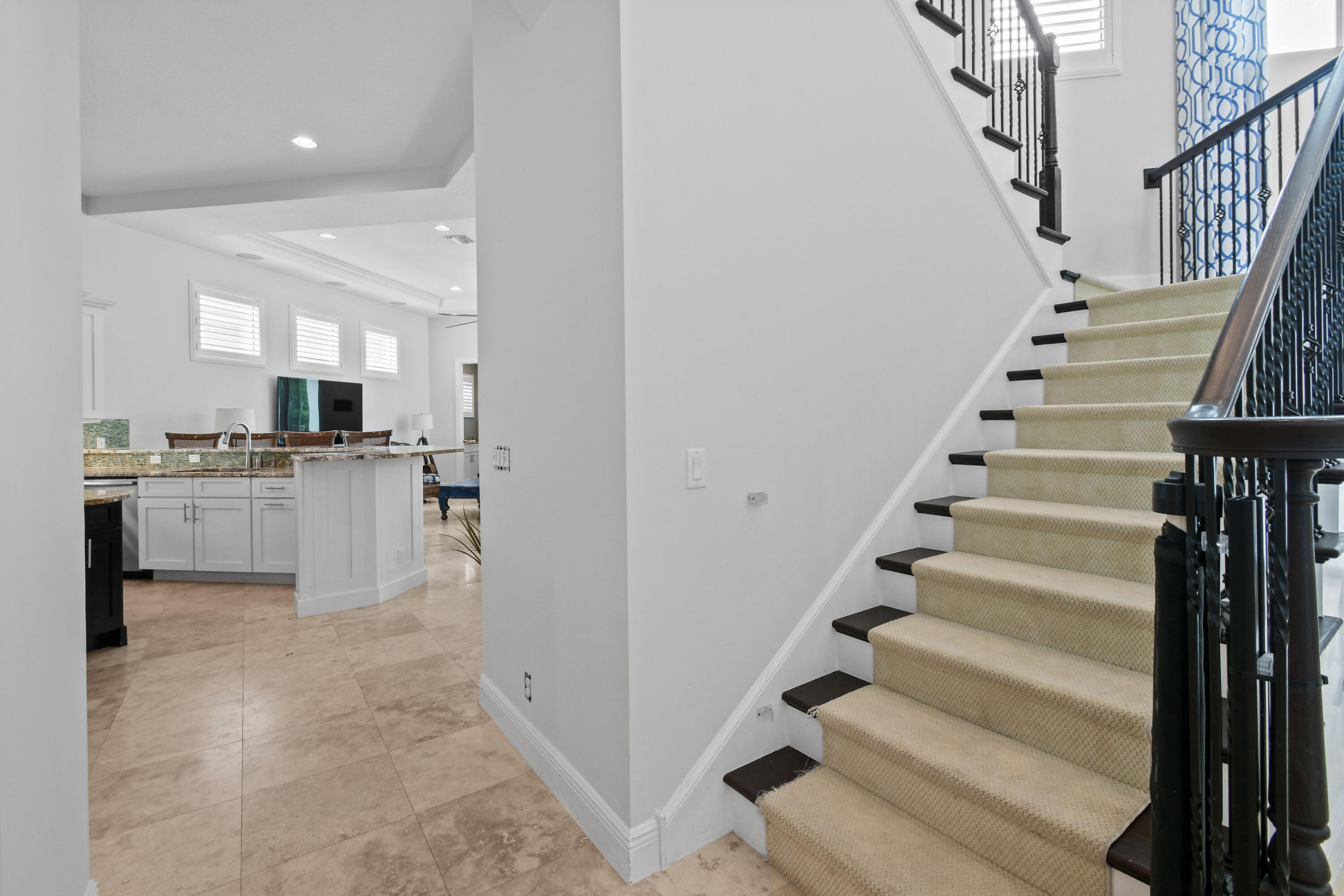 Carpeted staircase with dark railings beside a bright kitchen with white cabinets and a granite island.