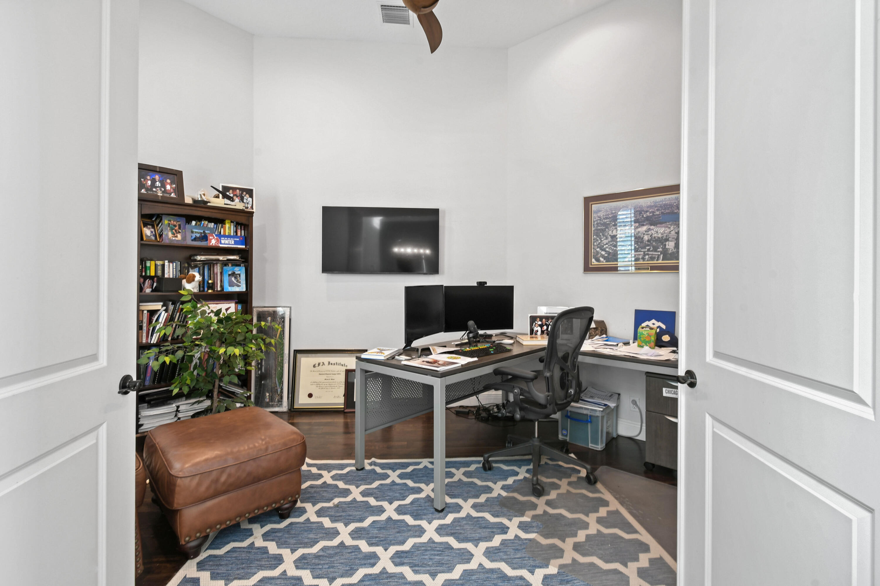 Furnished home office with an L-shaped desk, dual monitors, bookcase, and blue geometric rug.