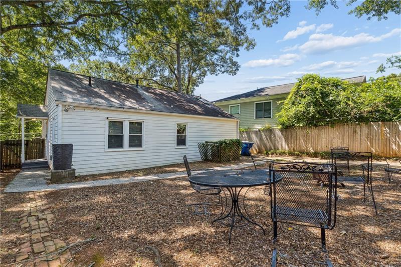 White house behind a backyard patio with a metal table and chairs beneath mature trees.