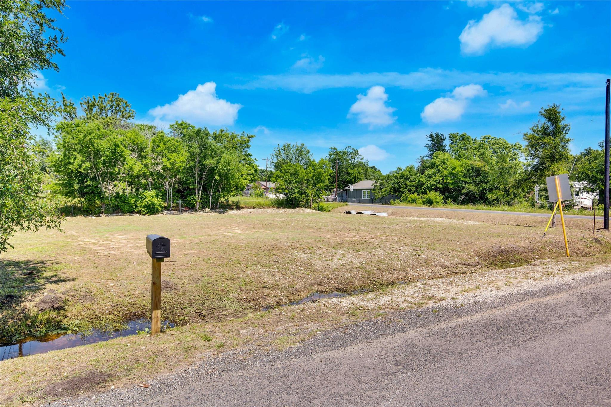 Cleared vacant lot with a roadside mailbox, gravel road, and trees along the back edge.