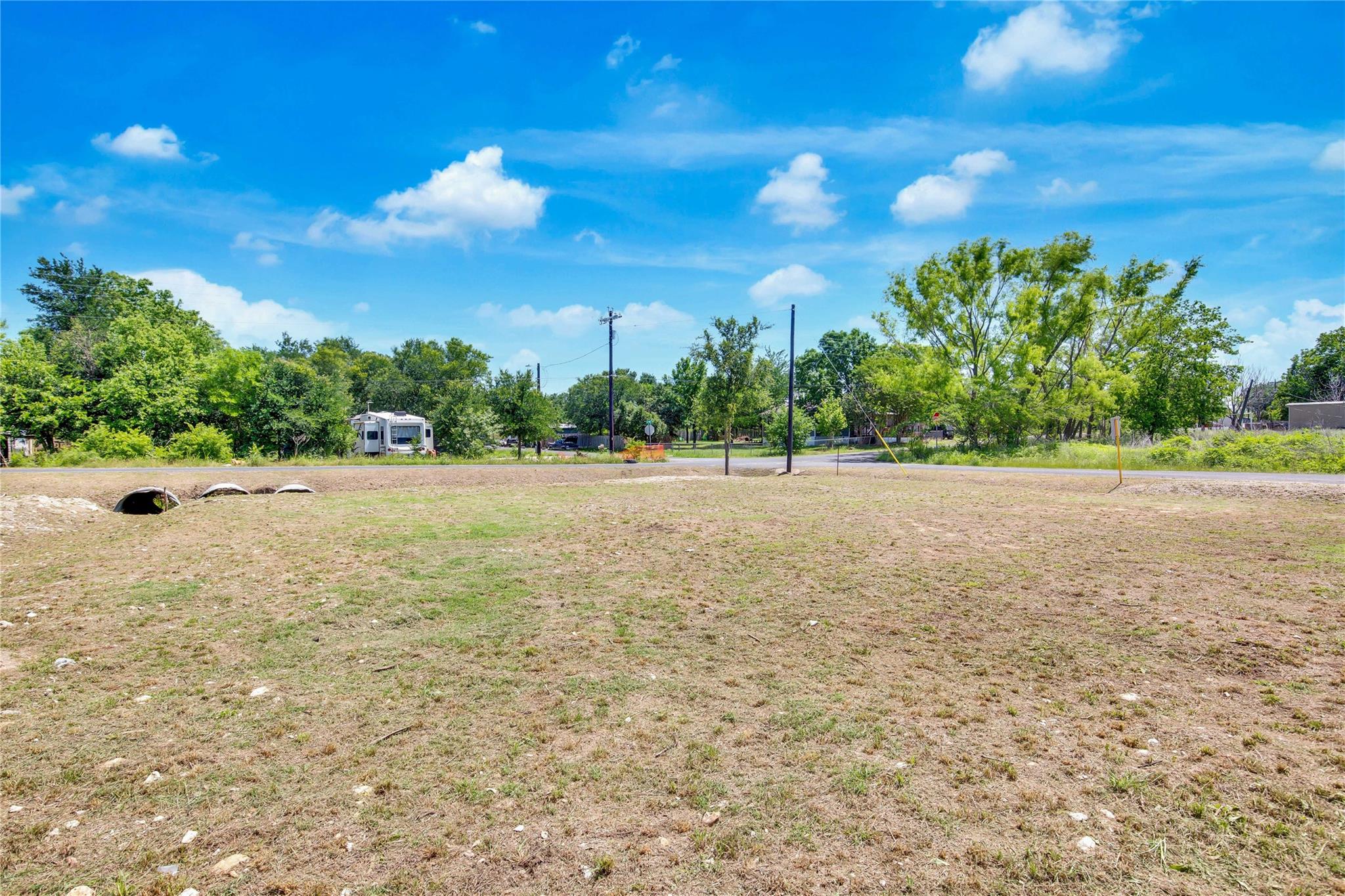 Vacant lot with dry grass, roadside utility poles, culvert pipes, and trees beyond.